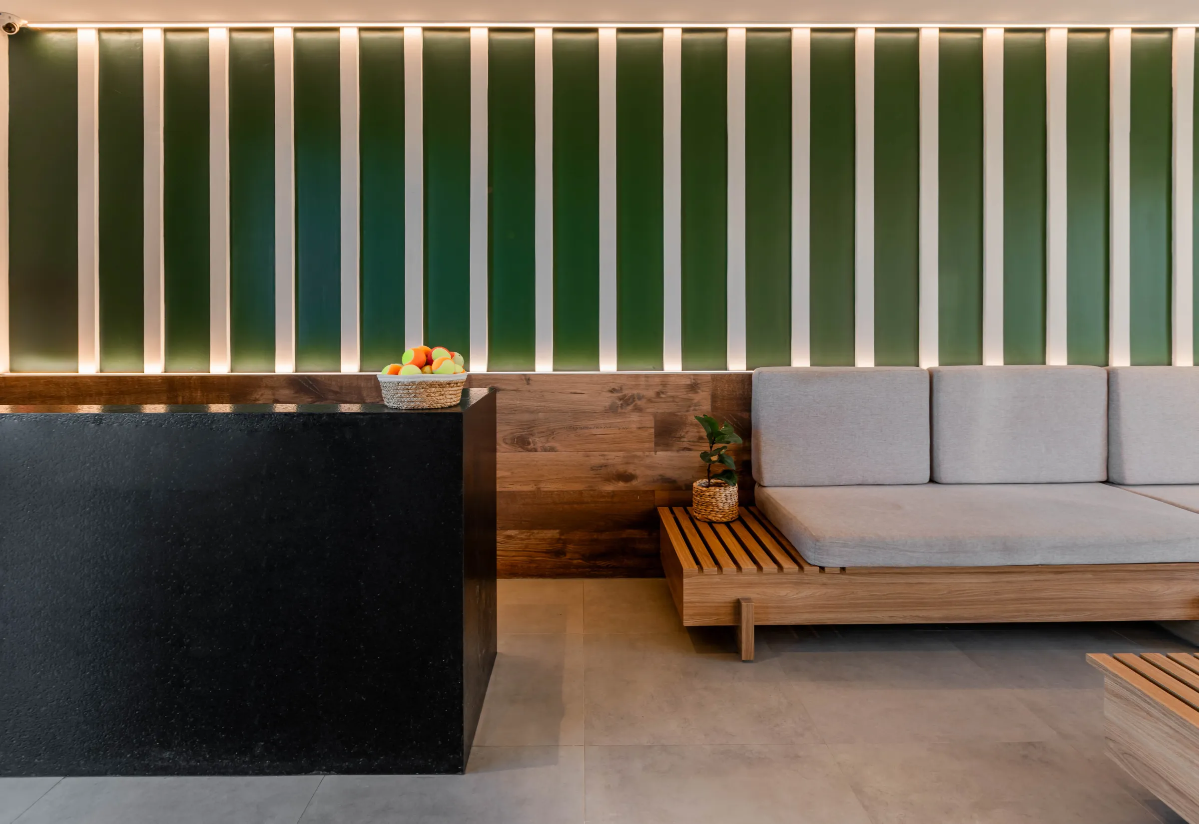 Lounge da Arena Carmel com parede verde ripada retroiluminada, balcão de granito preto com cesto de bolinhas de beach tennis e sofá de madeira com almofadas cinza