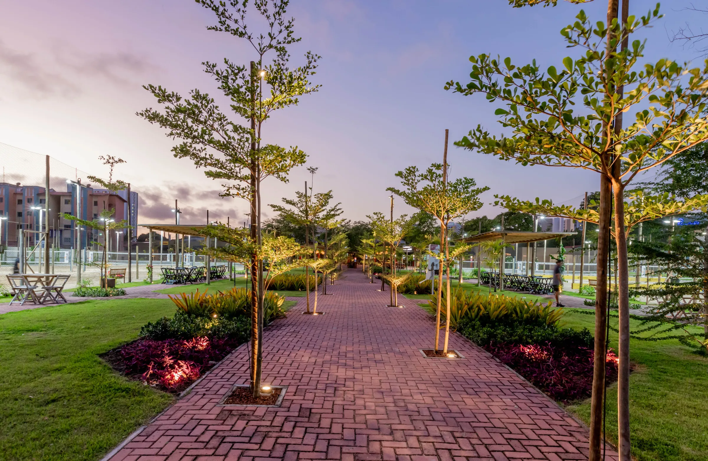 Praça arborizada da Arena Carmel ao entardecer com caminho de bloco intertravado ladeado por árvores iluminadas, canteiros floridos e mesas sob pergolados