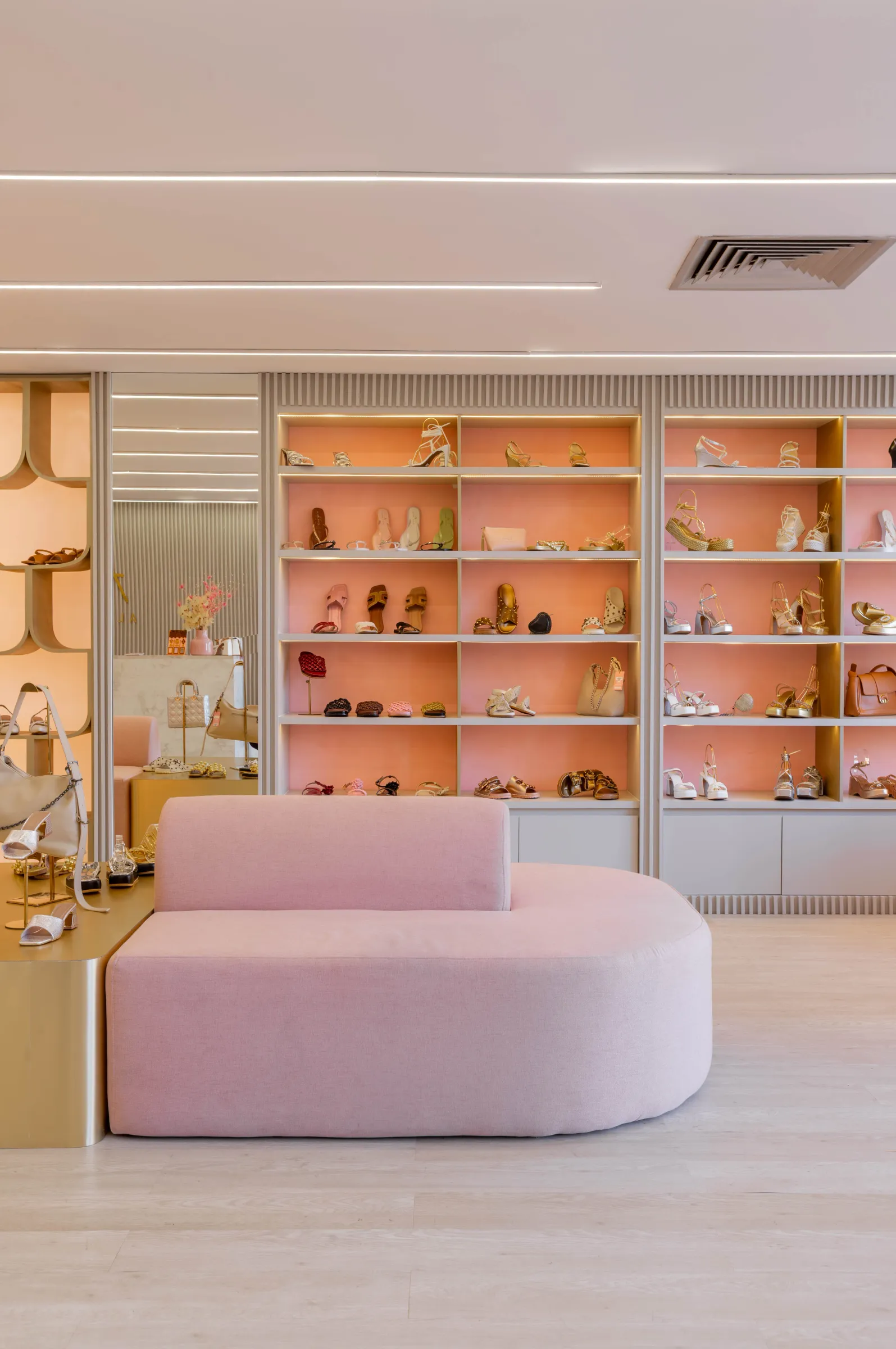 Interior da loja de calçados AV by Aline Vieira com estante iluminada de sapatos e sandálias com fundo rosa, banco curvo rosa ao centro e vitrine escultural dourada com nichos arredondados à esquerda