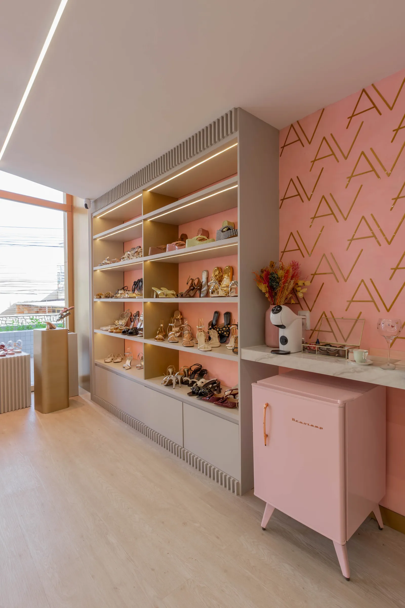 Ambiente da loja AV by Aline Vieira com estante de calçados iluminada, parede salmão com monograma AV dourado, frigobar retrô rosa e bancada de mármore com máquina de café e arranjo de flores secas