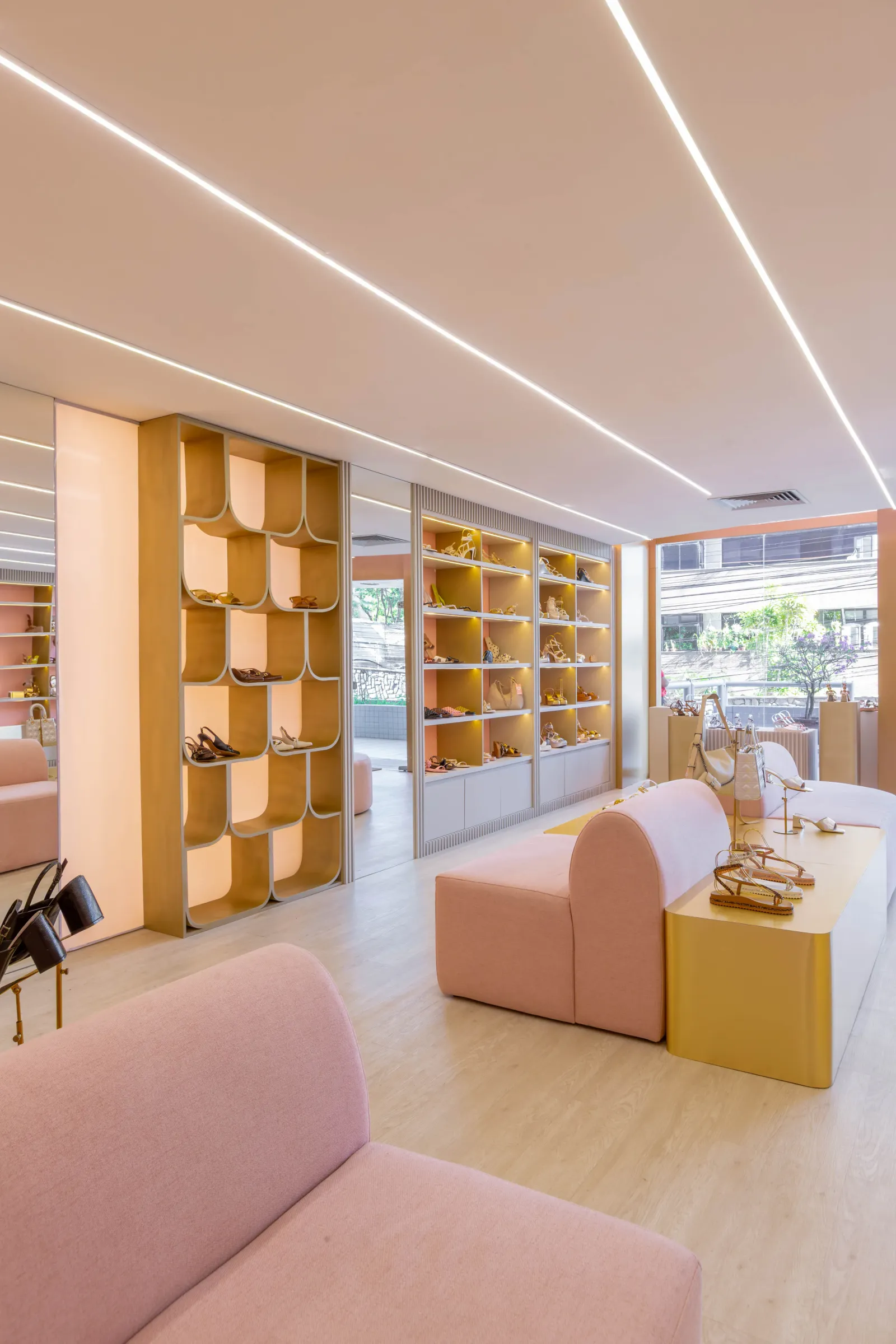 Salão da loja AV by Aline Vieira com estantes de sapatos e bolsas iluminadas, vitrine dourada de nichos curvos, bancos rosa e mesa expositora dourada com sandálias sob luz linear no teto