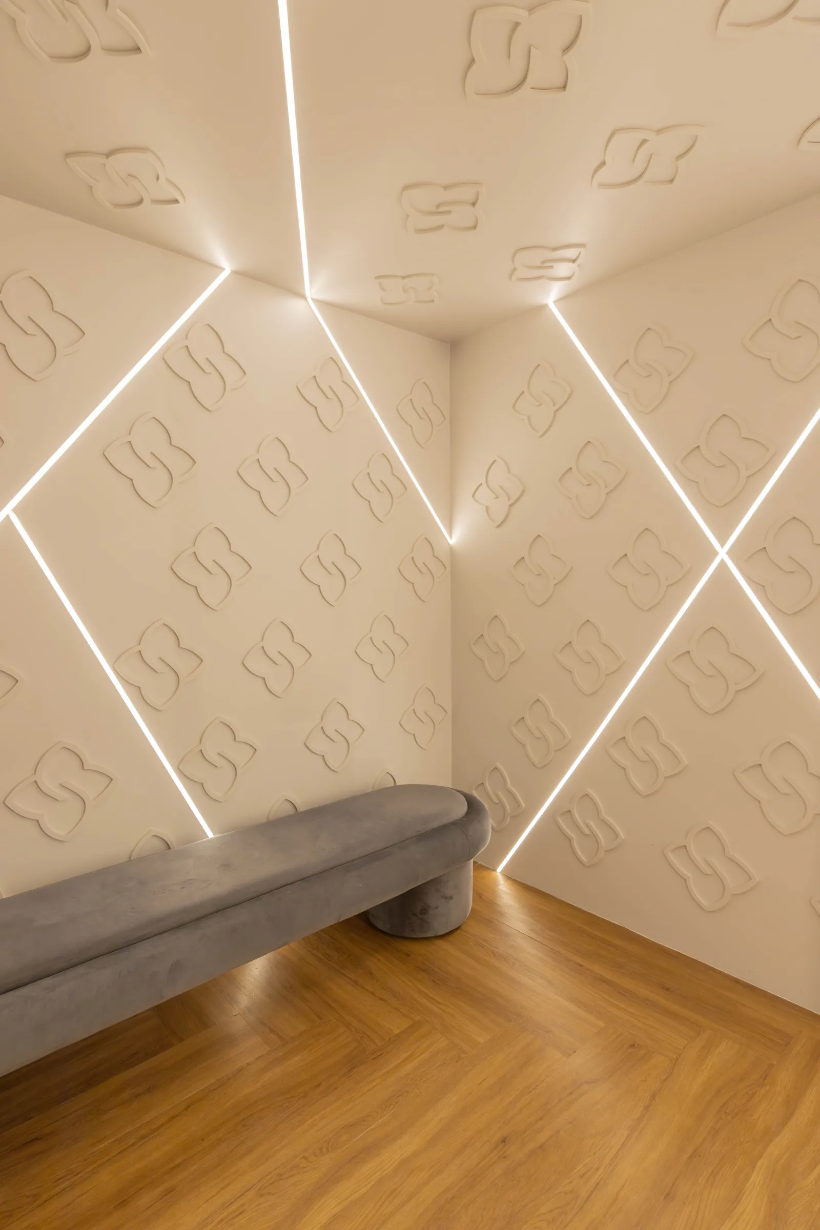 Detalhe de parede da boutique Donna Evellyn com painel de relevo do monograma da marca em tom off-white, iluminação de LED em linhas diagonais e banco curvo cinza sobre piso de madeira em espinha de peixe