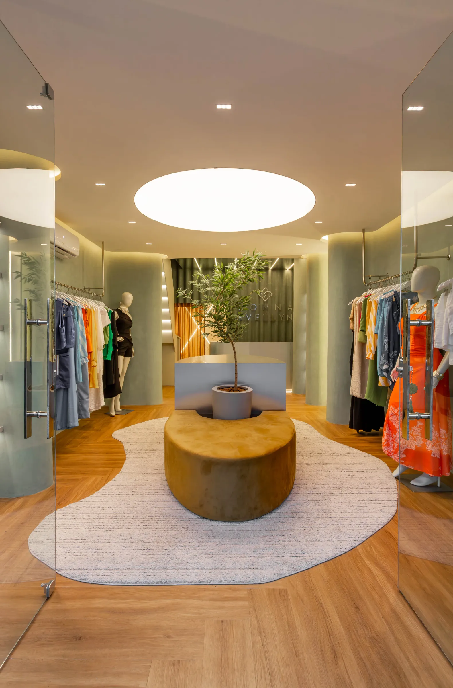 Salão da boutique Donna Evellyn com claraboia oval iluminada no teto, ilha central com oliveira em vaso, banco curvo de suede mostarda sobre tapete orgânico, araras com roupas coloridas e paredes de suede verde-sálvia
