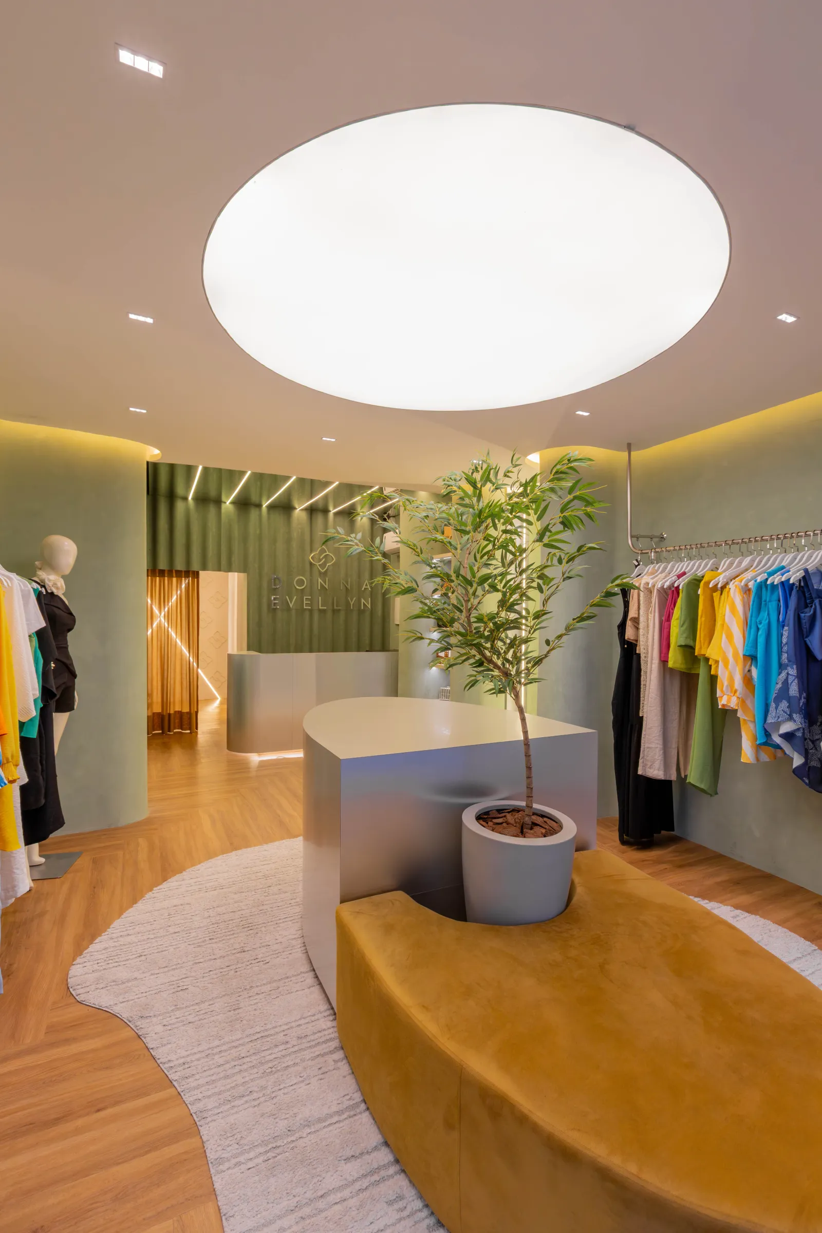 Vista do salão da boutique Donna Evellyn com logotipo da marca em parede canelada verde, oliveira central em vaso claro, banco curvo mostarda, araras de roupas coloridas e claraboia oval iluminada