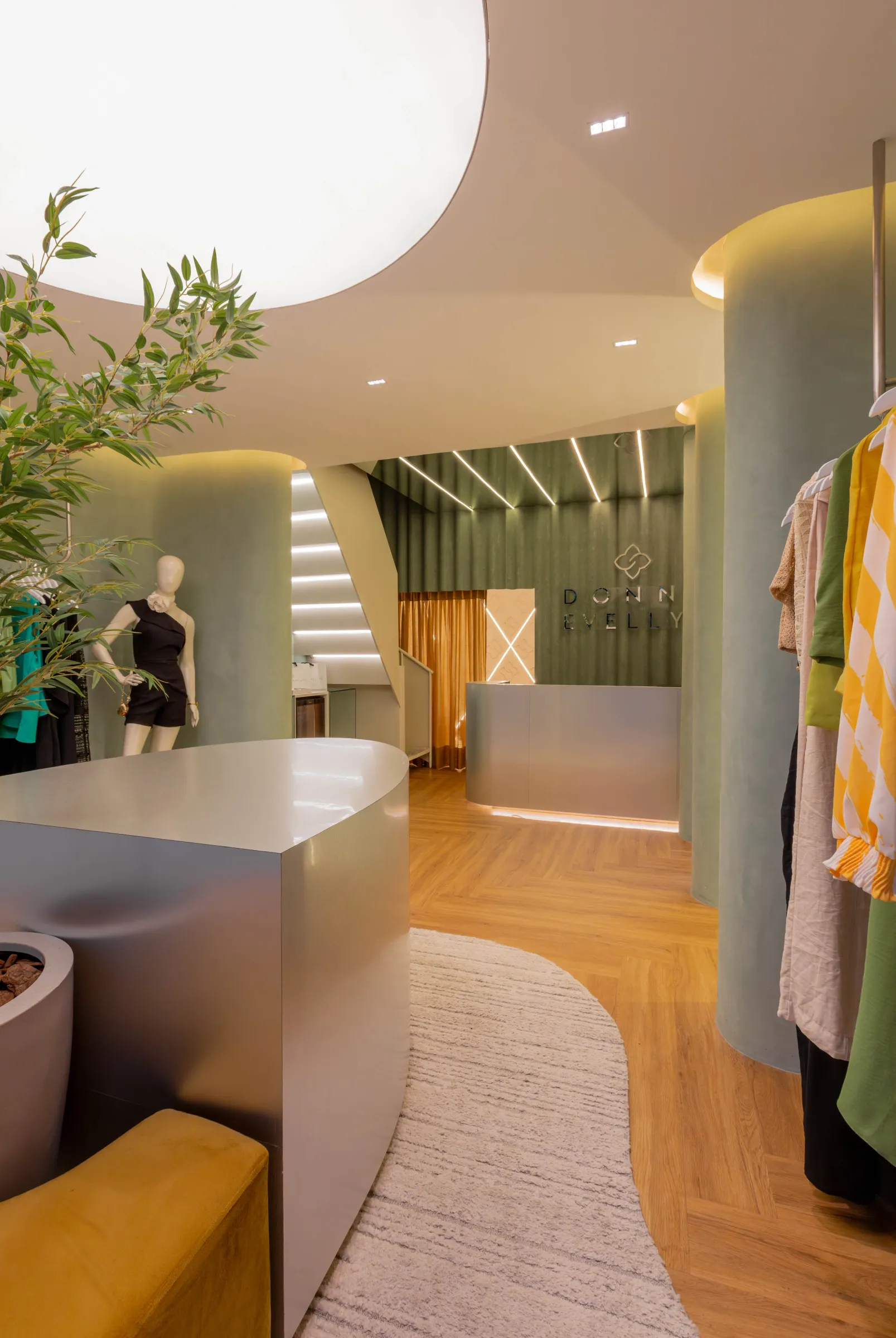 Ângulo da boutique Donna Evellyn com balcão curvo de aço escovado, manequim em macacão preto, colunas revestidas de suede verde e escada com iluminação linear ao fundo