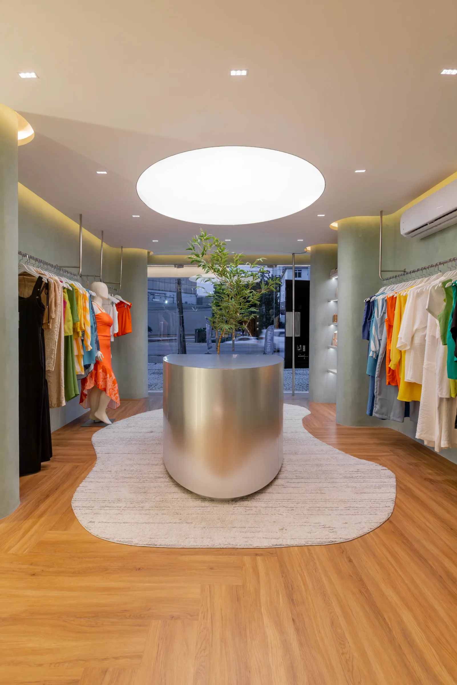 Vista ampla do salão da boutique Donna Evellyn com balcão cilíndrico de aço escovado ao centro, oliveira, claraboia oval, vitrine de vidro voltada para a rua e araras de roupas coloridas nas laterais