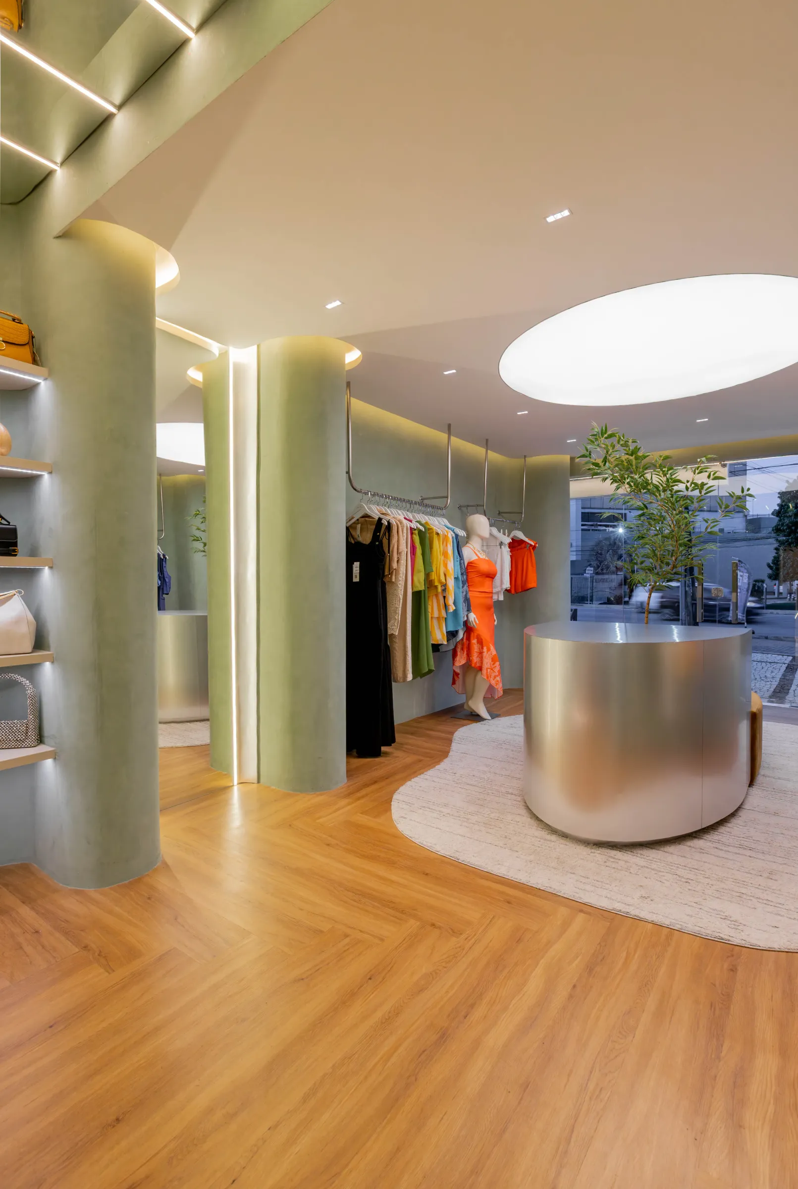 Ângulo da boutique Donna Evellyn com colunas curvas de suede verde, prateleiras com bolsas iluminadas, manequim em vestido laranja e balcão cilíndrico de aço escovado próximo à vitrine