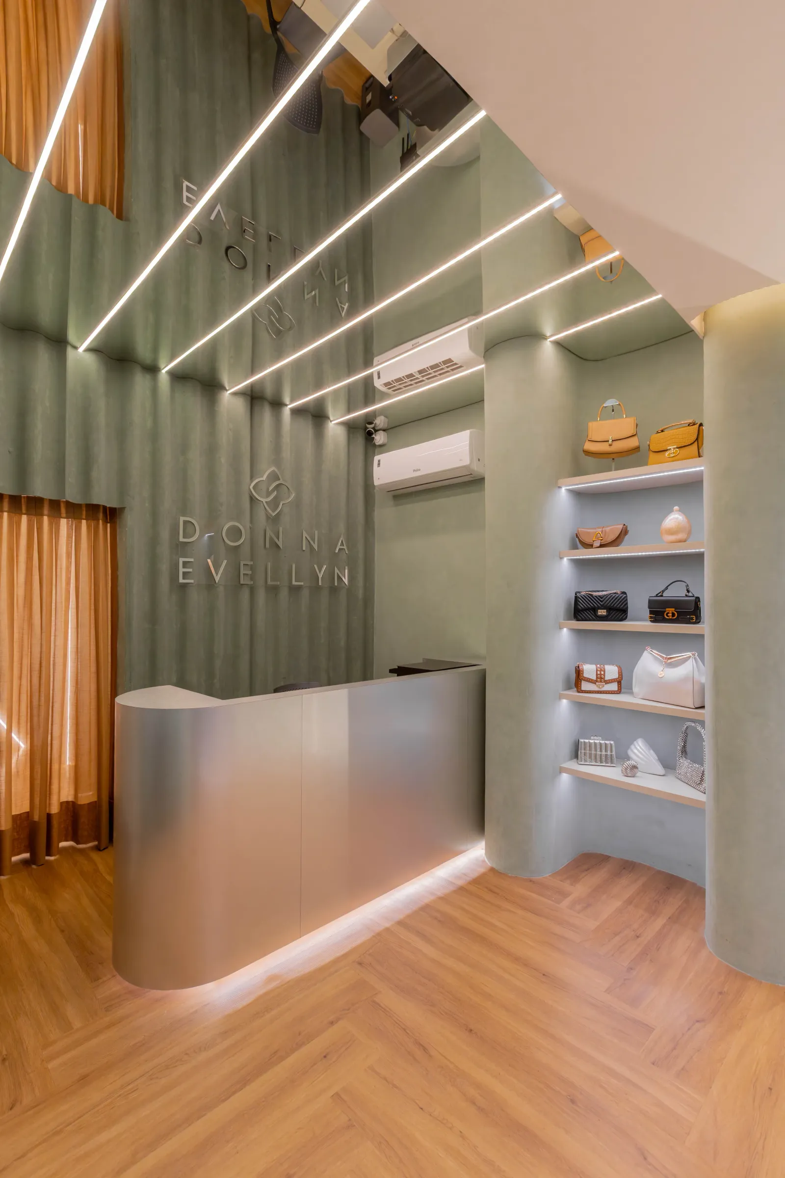 Área de caixa da boutique Donna Evellyn com balcão curvo de aço escovado, logotipo da marca em parede canelada verde, prateleiras com bolsas e cortinas em tom caramelo