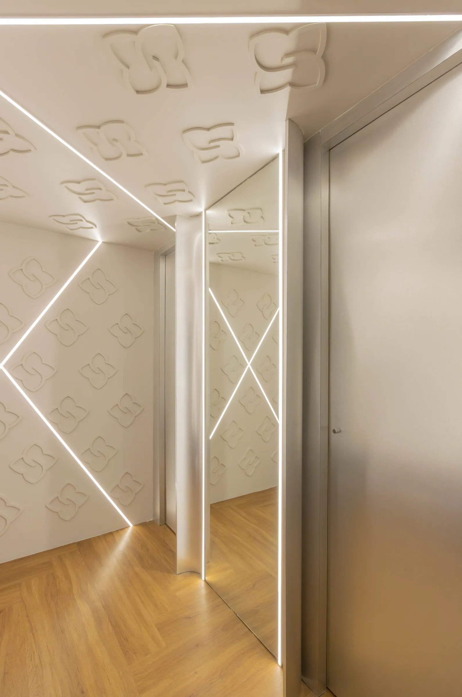 Corredor de provadores da boutique Donna Evellyn com paredes em relevo do monograma da marca, linhas de LED em desenho geométrico, porta espelhada e piso de madeira em espinha de peixe