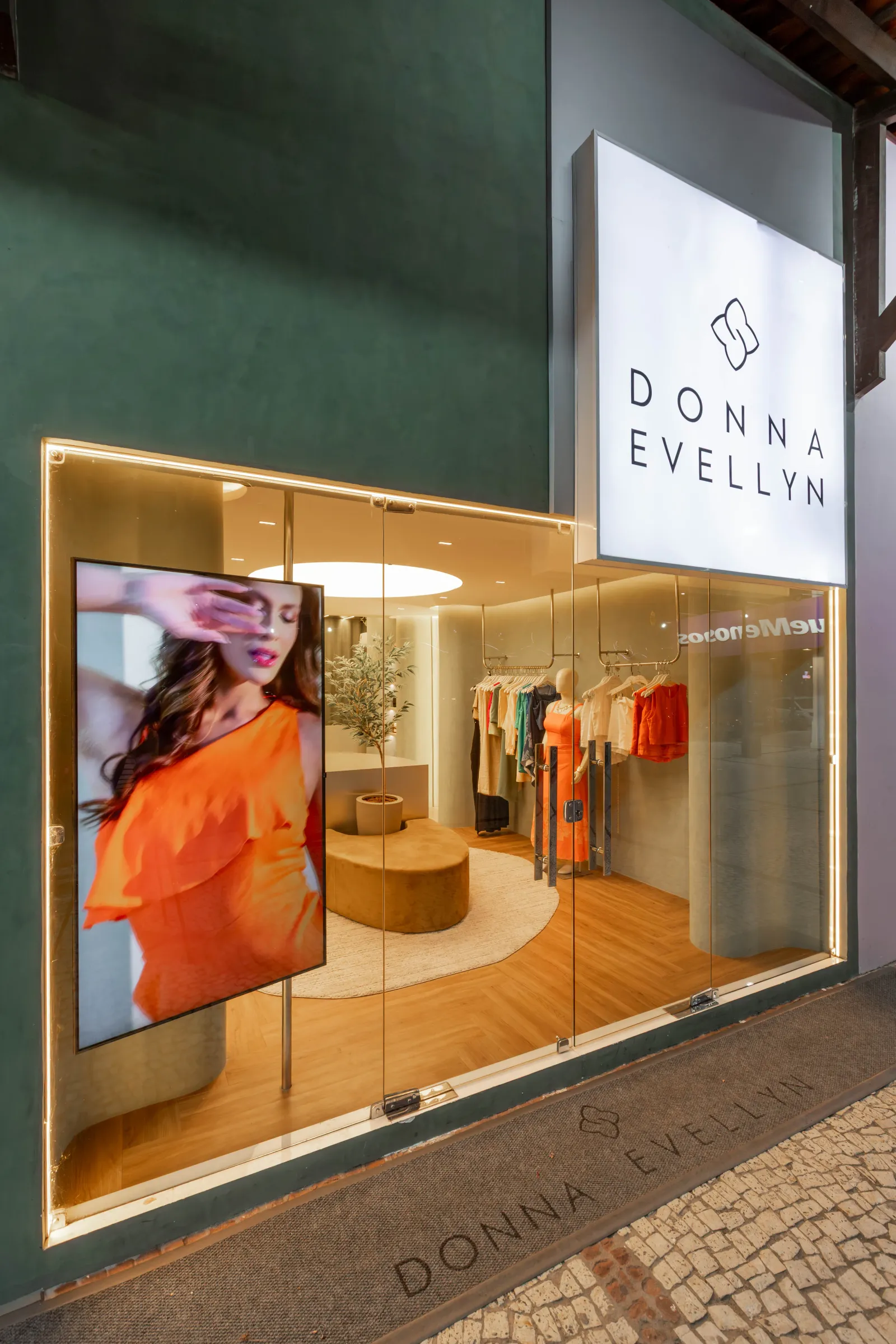 Fachada noturna da boutique Donna Evellyn com vitrine de vidro iluminada, letreiro luminoso da marca, tela digital com editorial de moda em laranja e capacho com o nome gravado na calçada