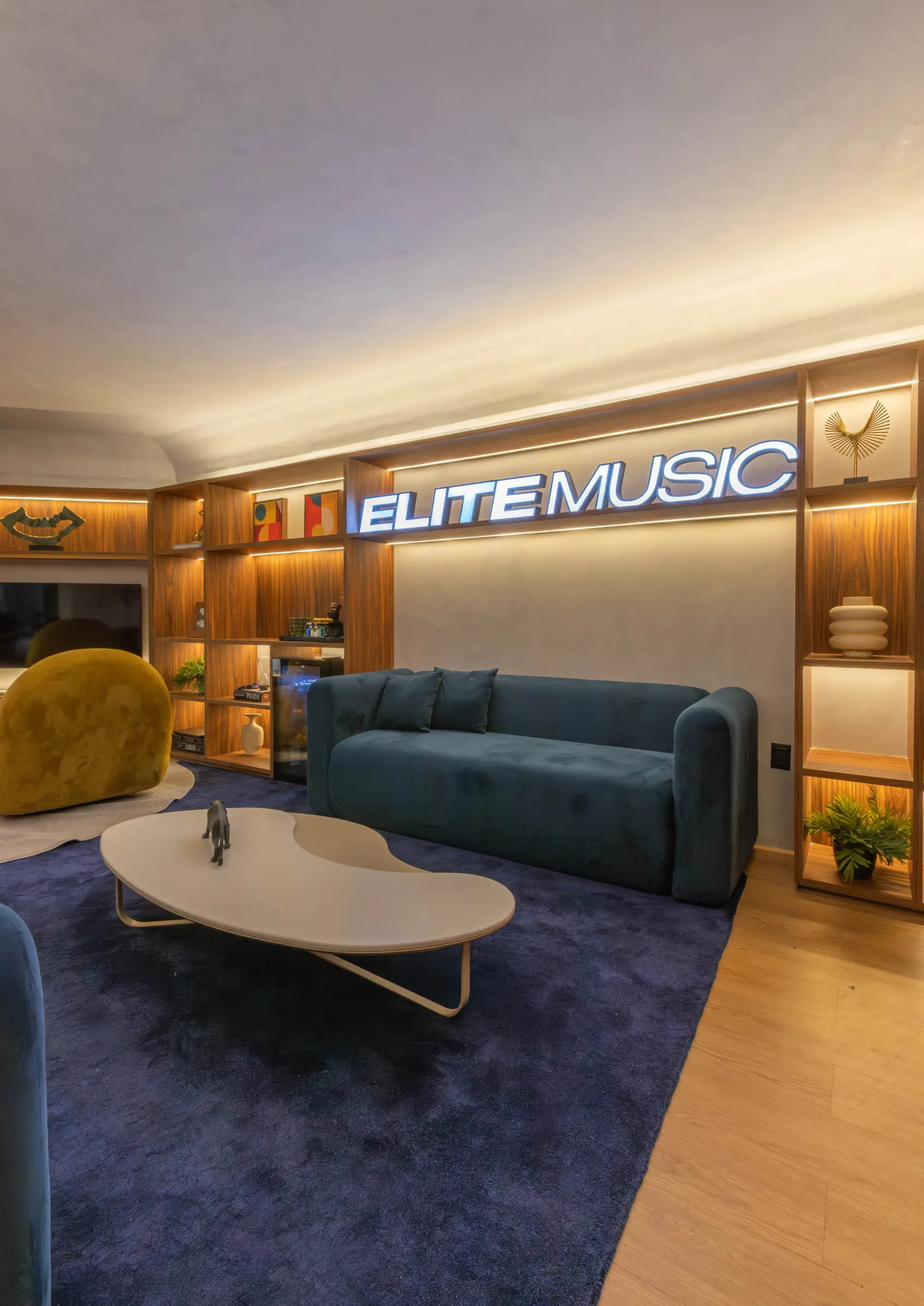 Lounge do escritório Elite Music com letreiro luminoso da marca em estante de madeira nobre com nichos iluminados, sofá de veludo verde, poltronas amarelo-mostarda, mesa de centro orgânica e tapete azul