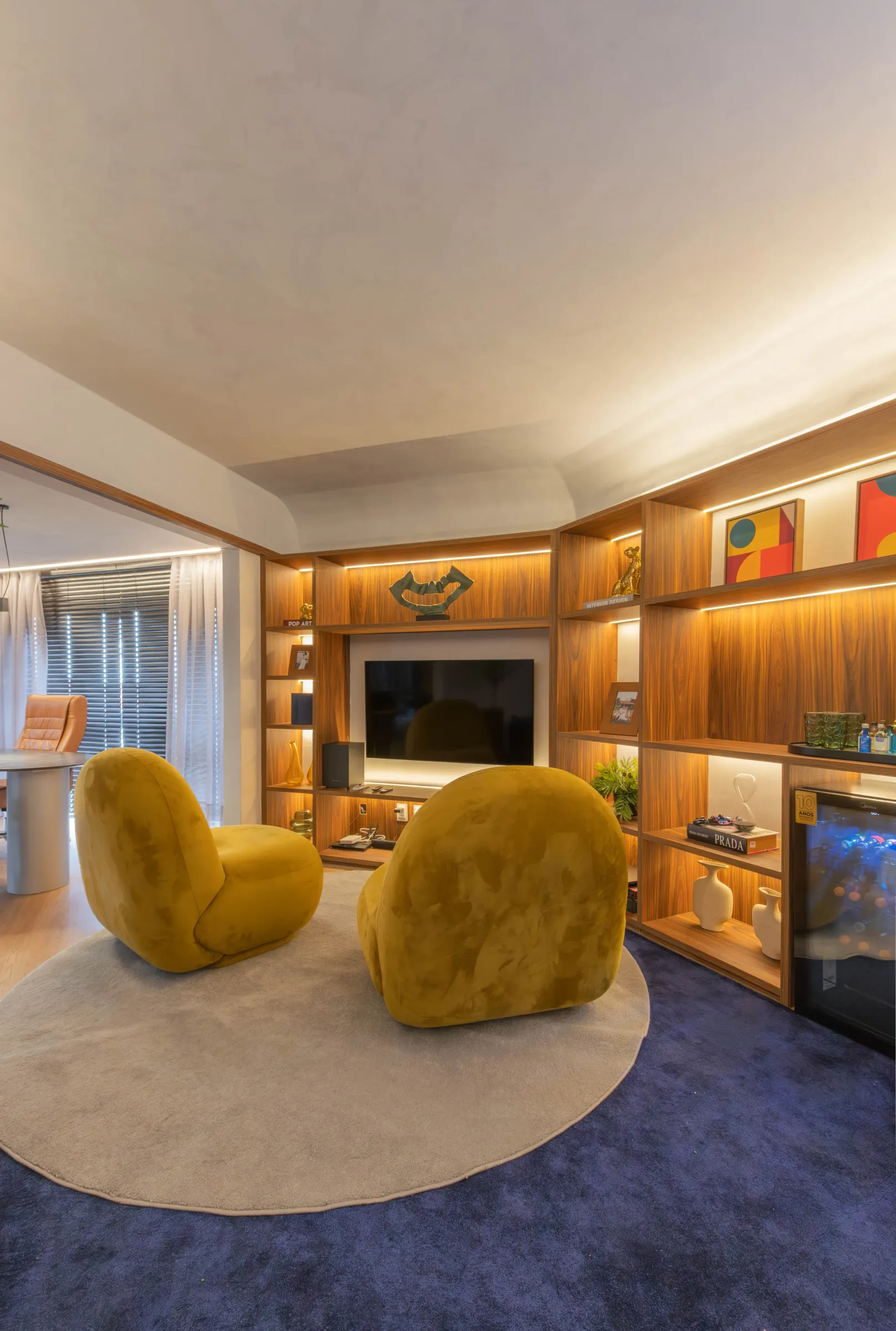 Estar do escritório Elite Music com duas poltronas escultóricas de veludo amarelo-mostarda sobre tapete redondo, painel de TV embutido em marcenaria de madeira com nichos iluminados e adega climatizada