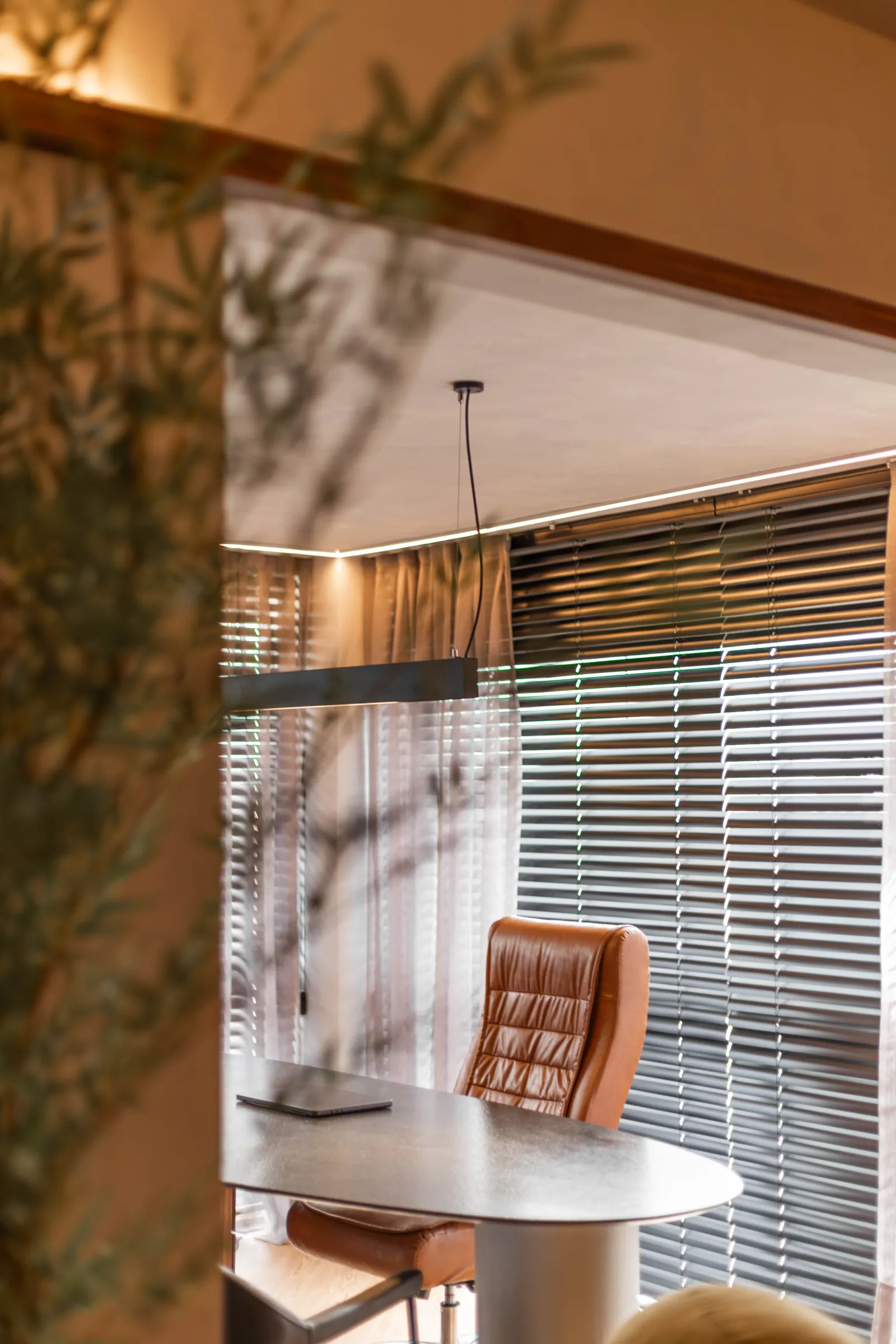 Mesa de trabalho do escritório Elite Music refletida em divisória de vidro, com poltrona de couro caramelo, cortinas e persianas de madeira filtrando a luz da janela