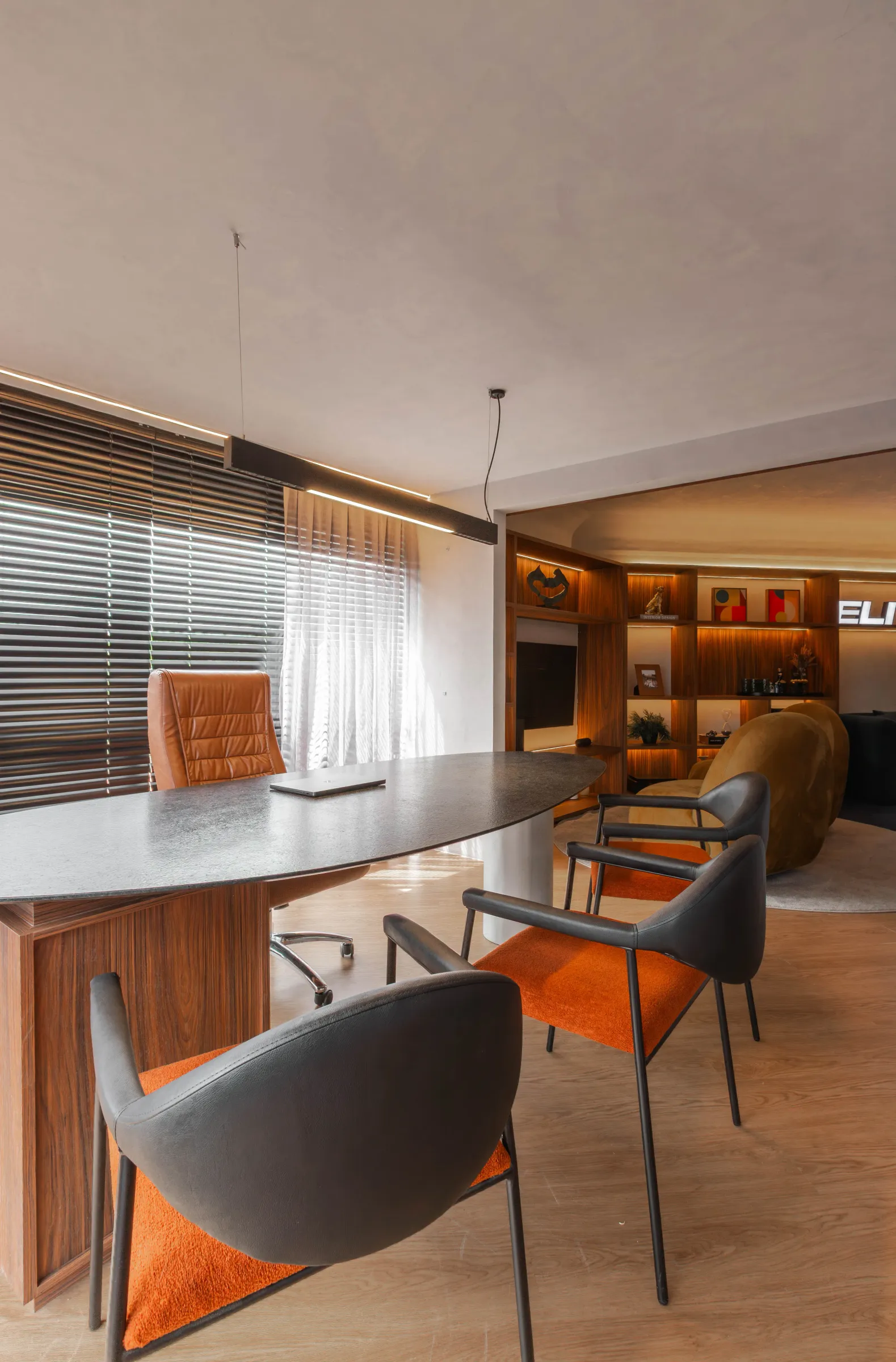 Mesa de reunião do escritório Elite Music com cadeiras de couro preto e assento laranja, cadeira presidente caramelo e o lounge com poltronas amarelas e letreiro da marca ao fundo