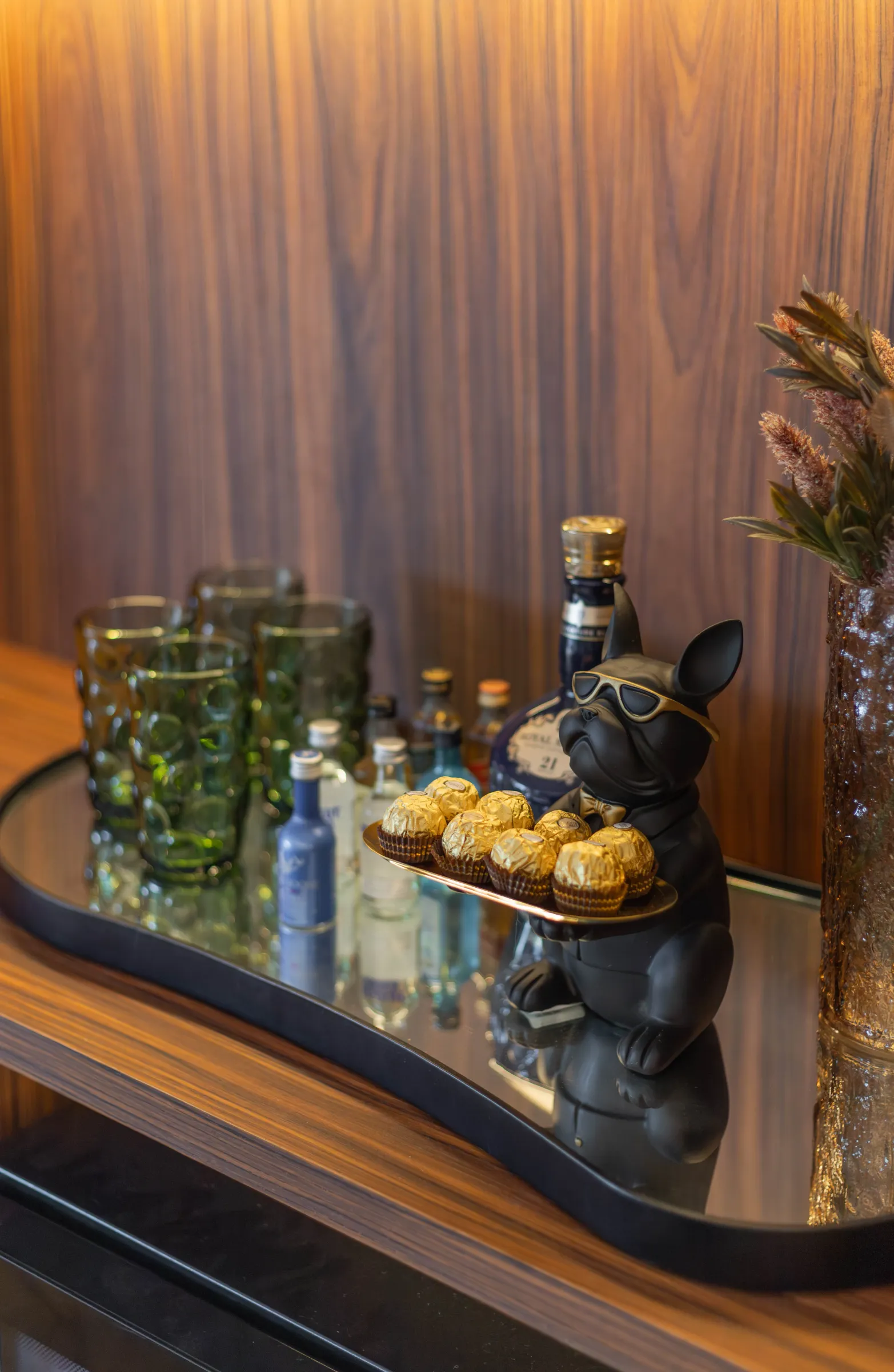 Detalhe decorativo do escritório Elite Music com bandeja espelhada sobre marcenaria de madeira exibindo copos verdes, miniaturas de bebida e estatueta de bulldog preto de óculos segurando bombons dourados