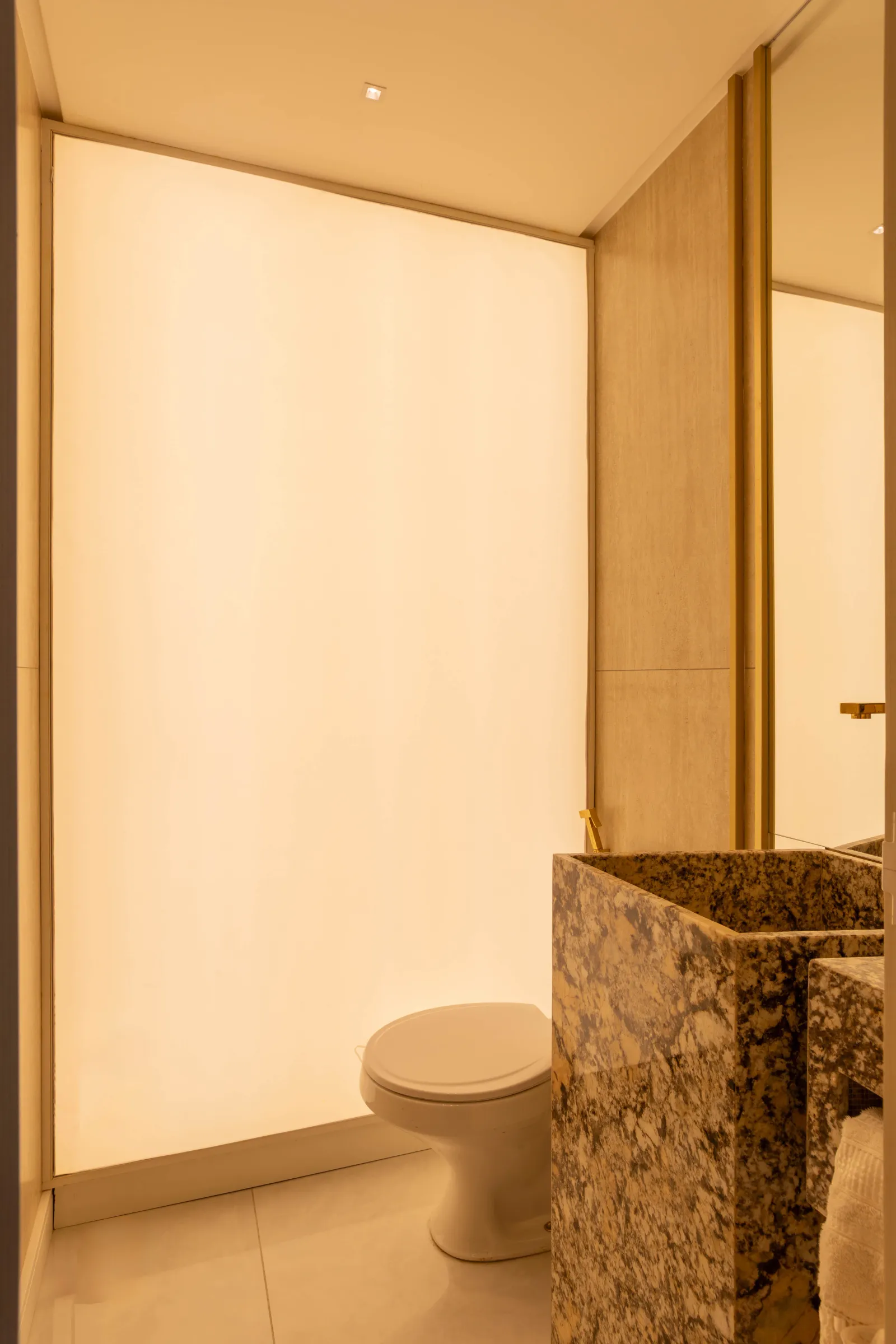 Lavabo da La Clinic com cuba sob medida esculpida em granito, torneira dourada de parede, painel de vidro retroiluminado ocupando a parede e espelho lateral