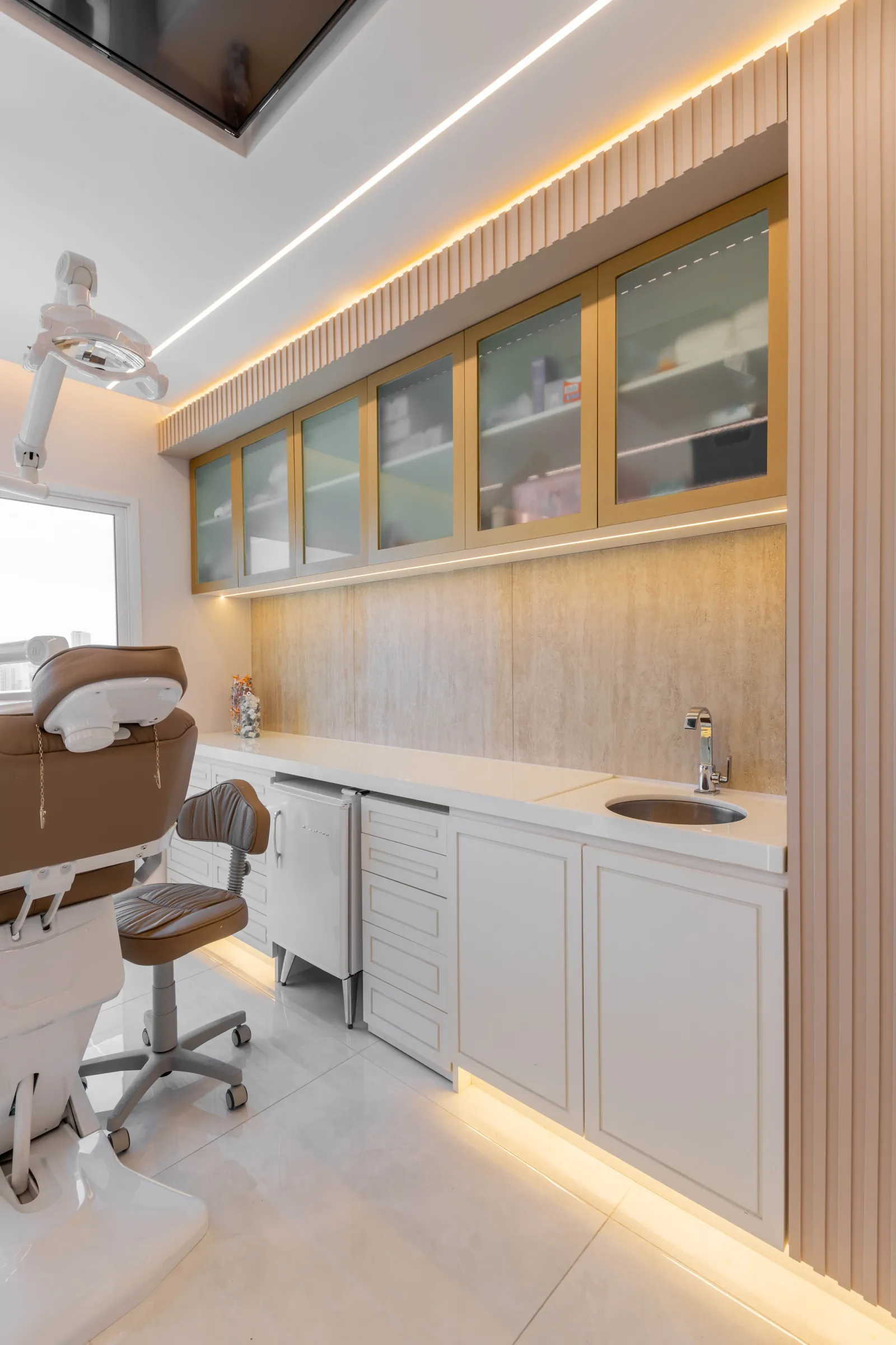 Sala de atendimento da La Clinic com cadeira odontológica/estética, refletor articulado, armários de vidro com moldura dourada e bancada branca com cuba sob iluminação indireta