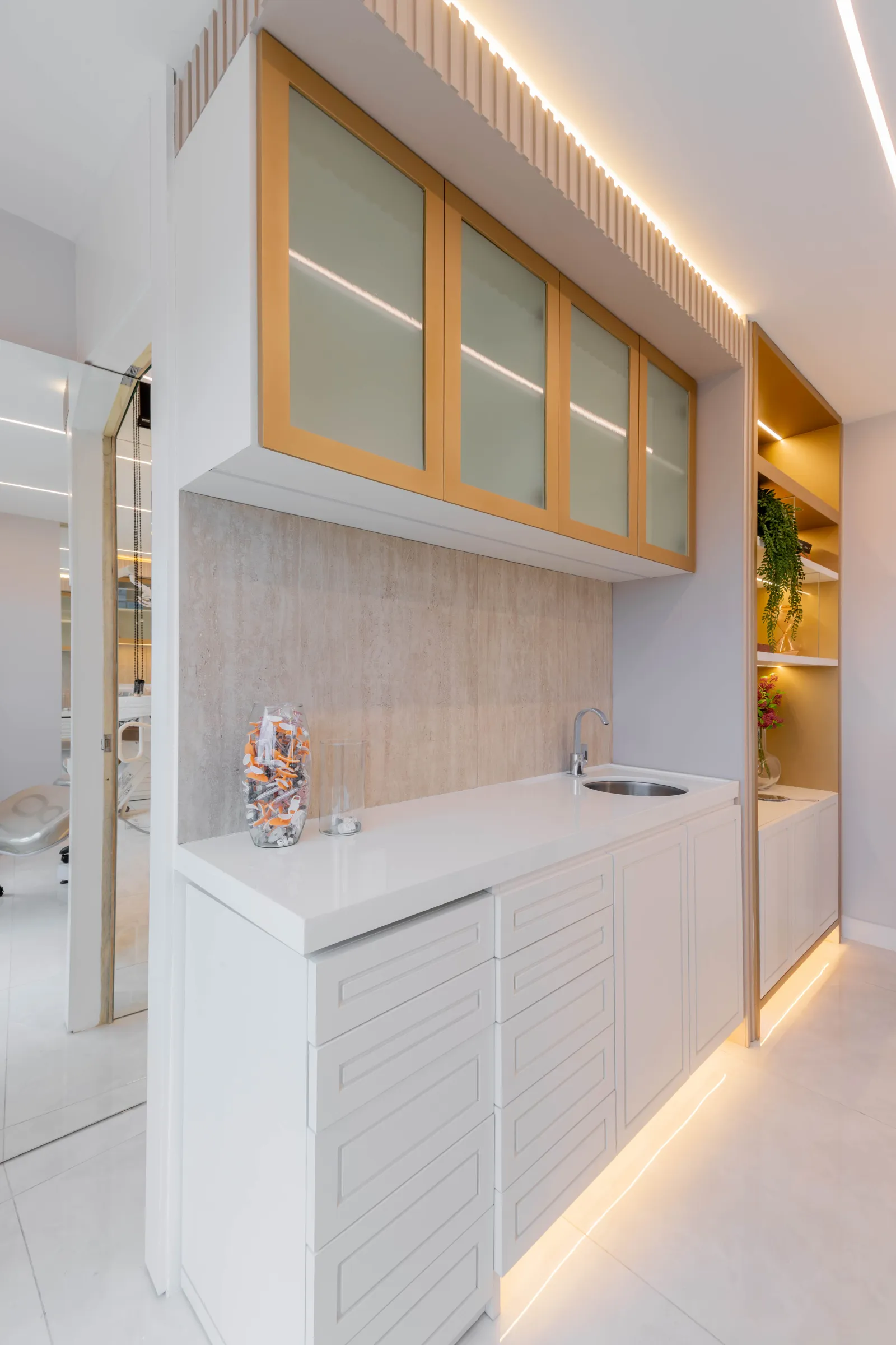 Bancada de apoio da La Clinic com armários brancos ripados, cuba e torneira, armário superior de vidro com moldura dourada e espelho refletindo a sala de procedimentos