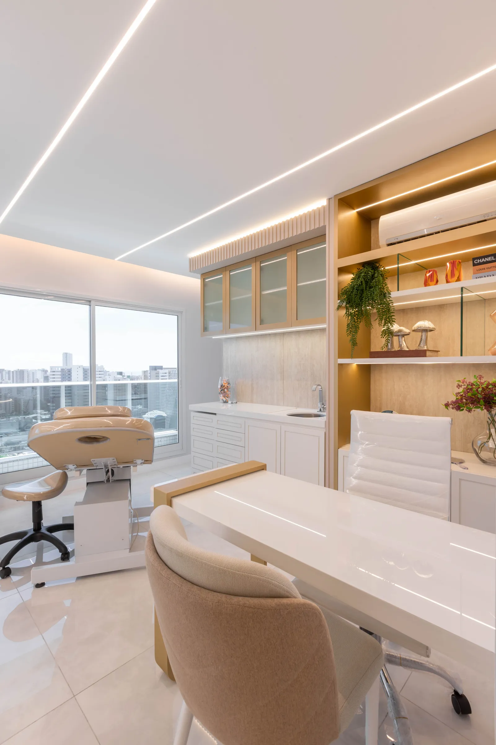 Consultório da La Clinic com maca de estética, mesa de atendimento branca e dourada, estante iluminada com livros de moda e objetos decorativos e janela ampla com vista da cidade