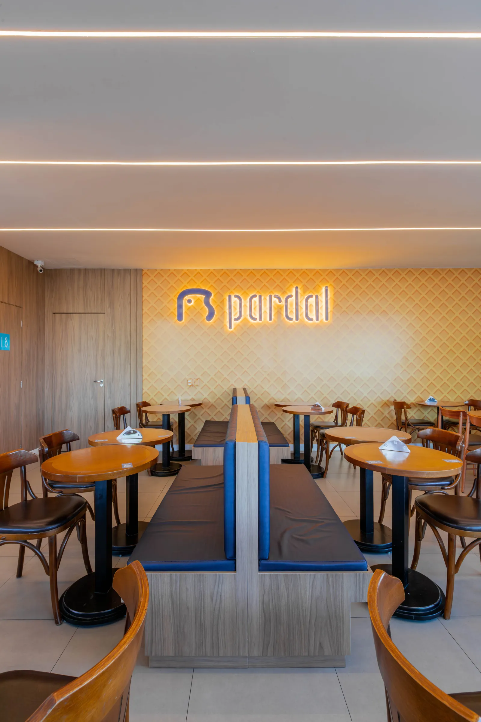 Salão da sorveteria Pardal com mesas redondas de tampo laranja, cadeiras de madeira estilo bistrô, banco central azul e parede texturizada de casquinha com o logo iluminado