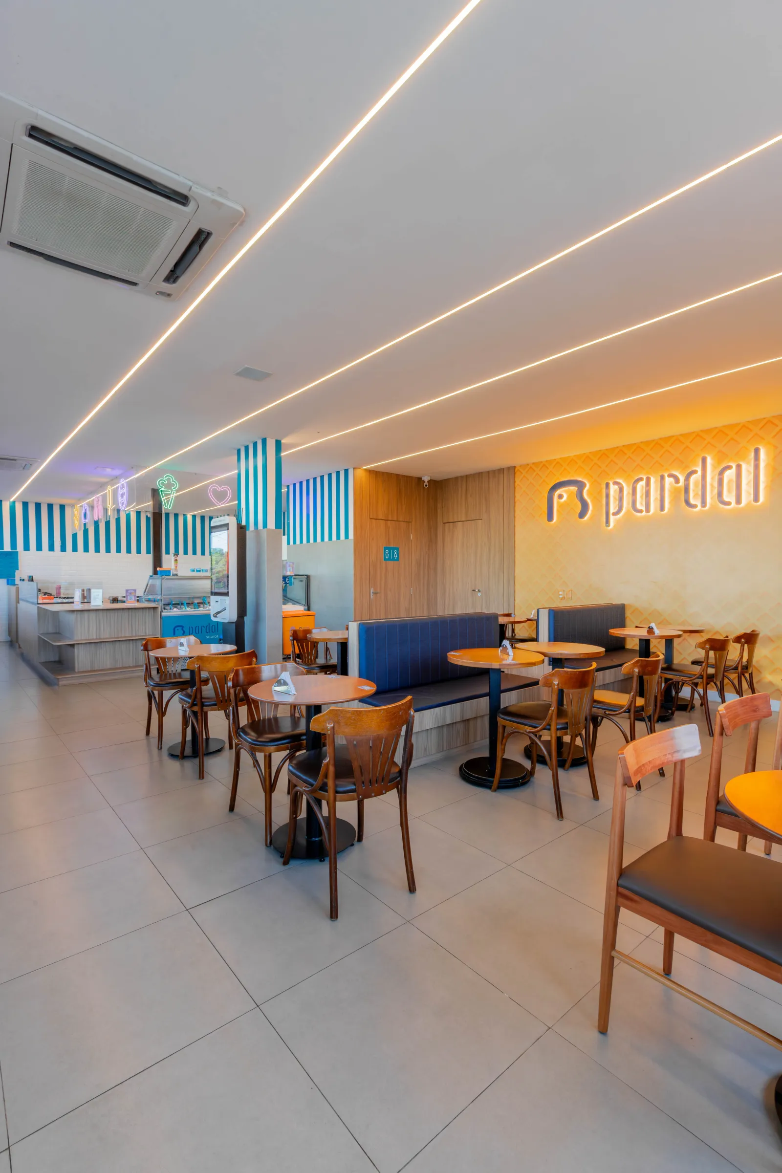 Salão da sorveteria Pardal com balcão de gelatos ao fundo, letreiros de neon, parede de listras azuis e mesas com bancos estofados azuis