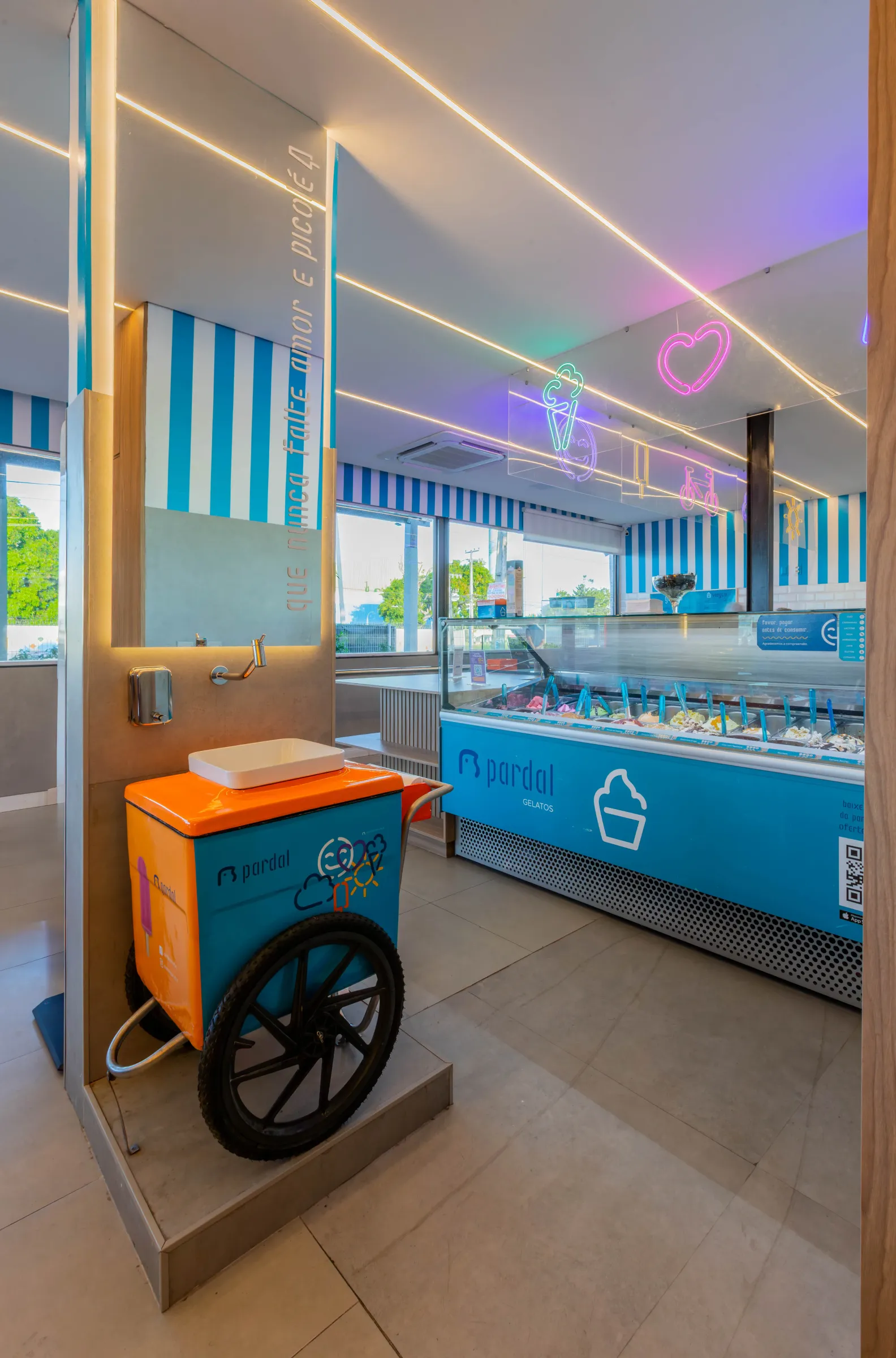 Balcão de gelatos coloridos da sorveteria Pardal, com carrinho de picolé da marca, letreiros de neon e parede de listras azuis
