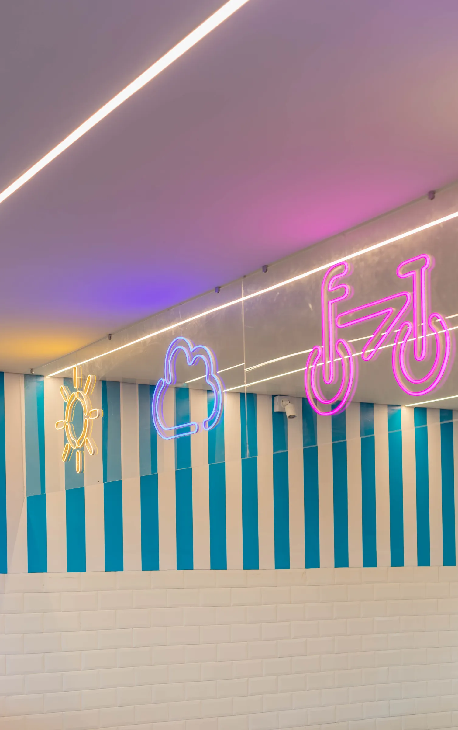 Parede da sorveteria Pardal com letreiros de neon de sol, nuvem e bicicleta, listras azul e branco e azulejo metrô branco