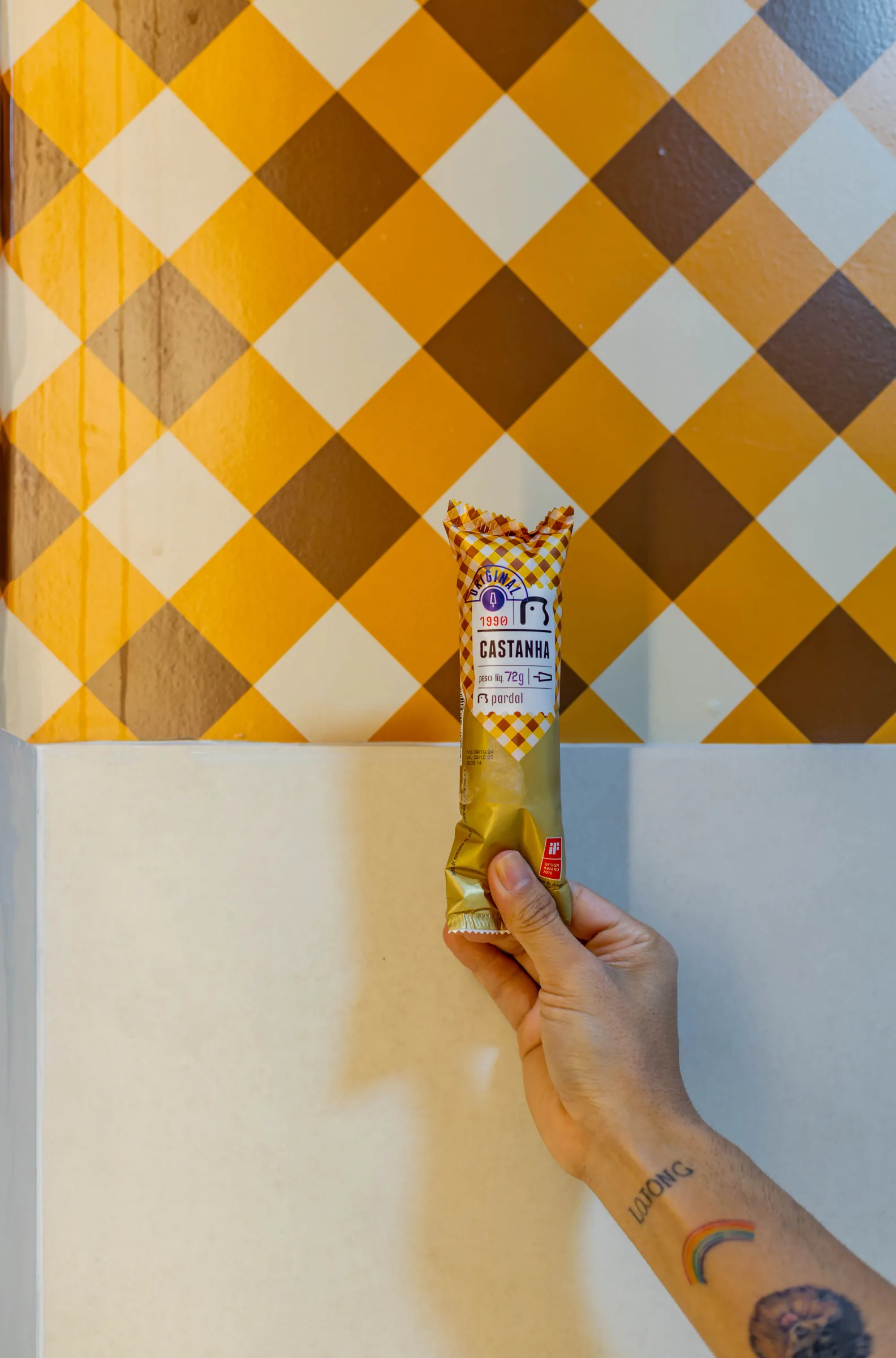 Mão segurando um picolé da Pardal sabor castanha embalado, diante de parede com padrão xadrez amarelo