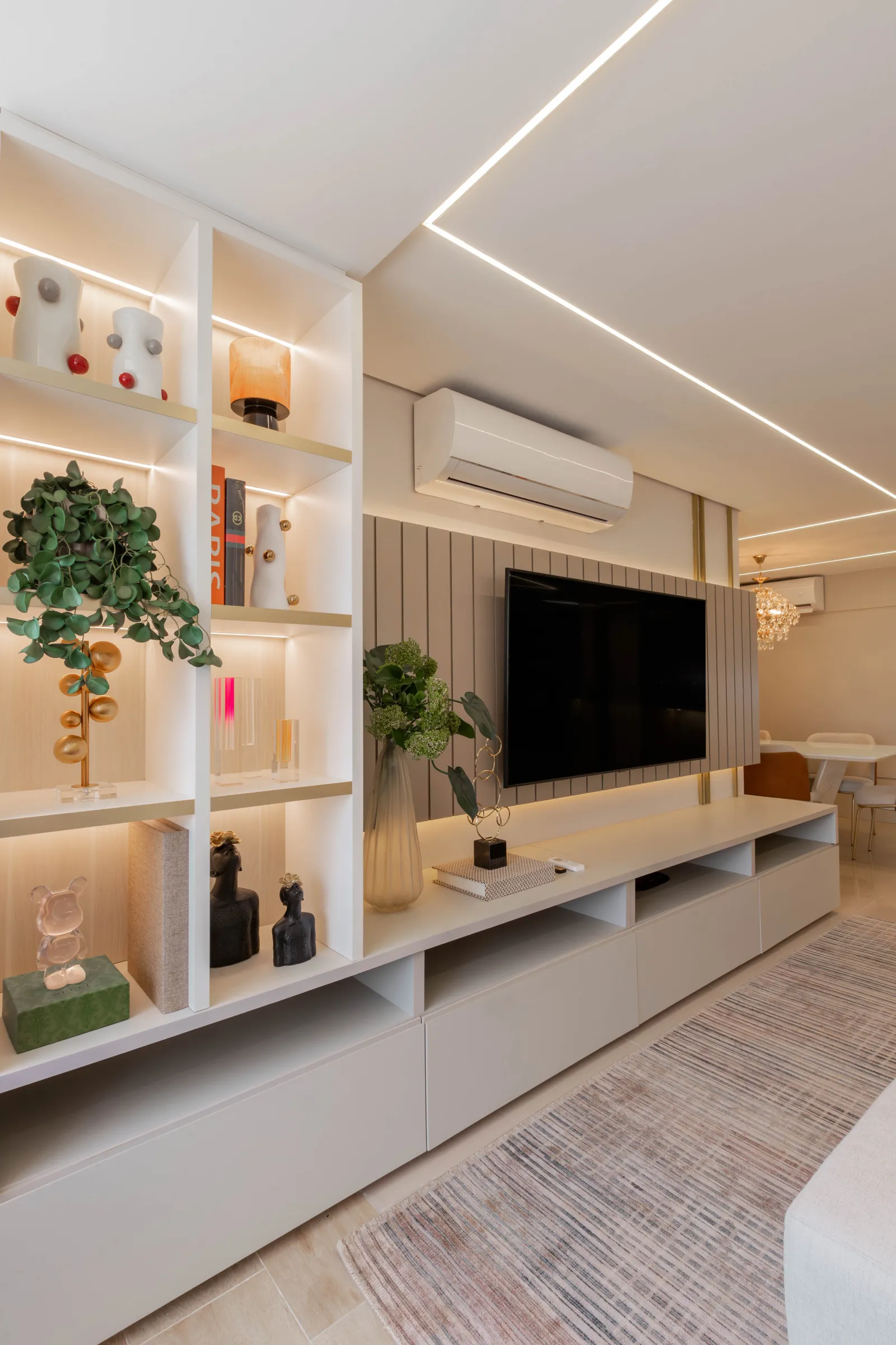 Sala de estar do Residencial LV com estante branca de nichos iluminados e objetos decorativos, painel ripado cinza com TV e rack branco, integrada à sala de jantar com lustre de cristal ao fundo