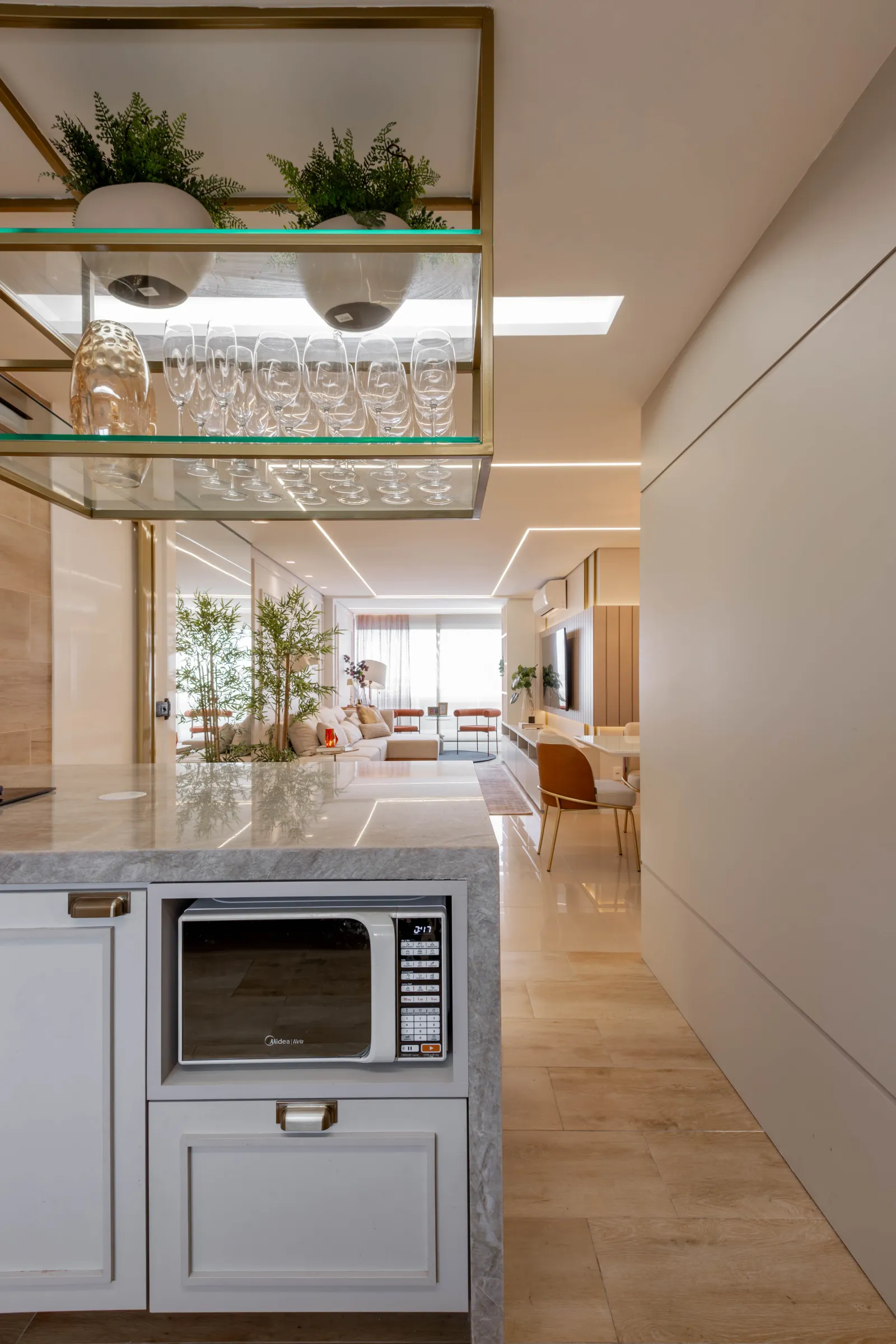 Detalhe da cozinha do Residencial LV com prateleira aérea dourada de taças e plantas, micro-ondas embutido na península de mármore e integração com o living ao fundo
