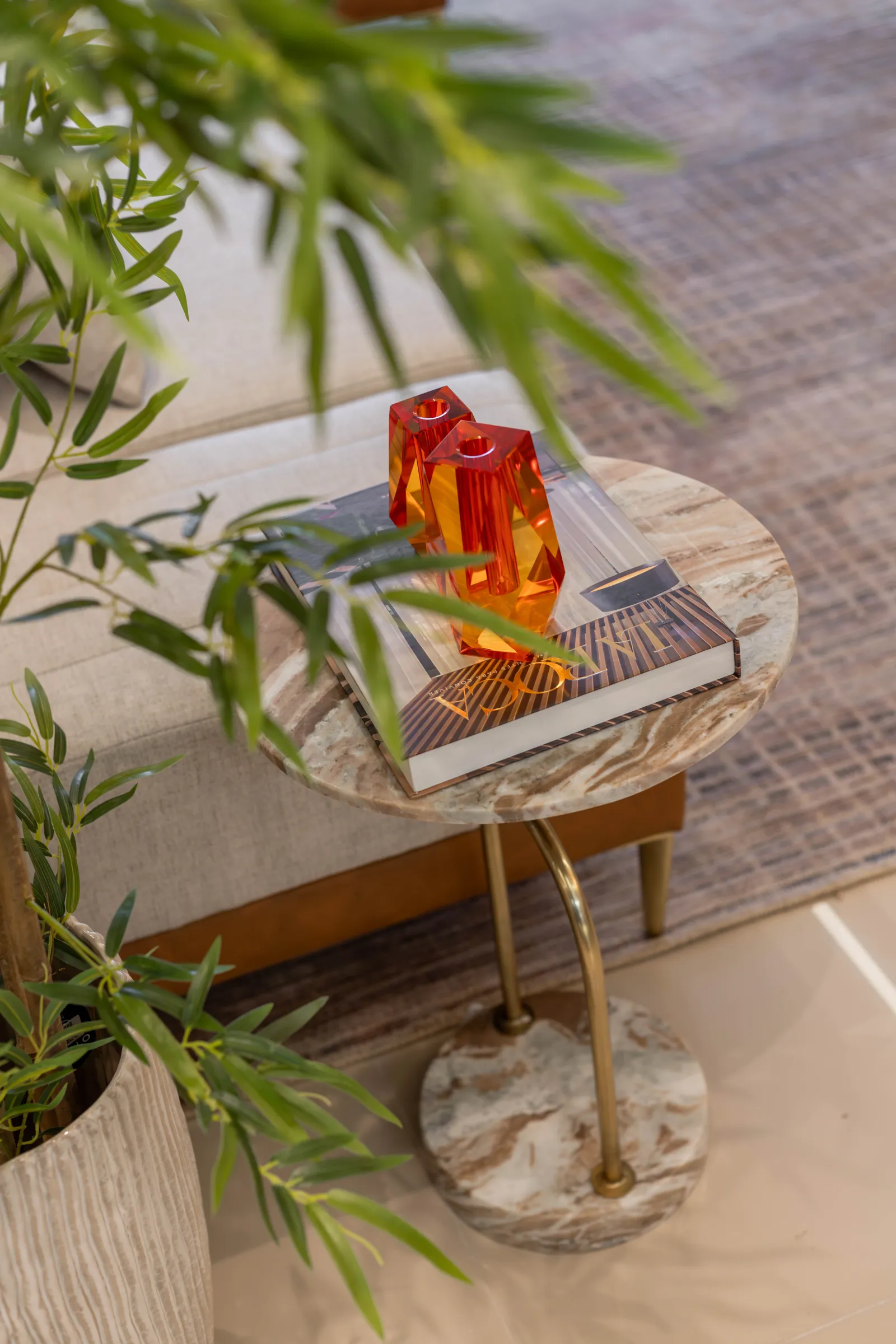 Detalhe decorativo do Residencial LV com mesa lateral de mármore, livro de capa dura e castiçais de cristal em tons de laranja, com planta em primeiro plano