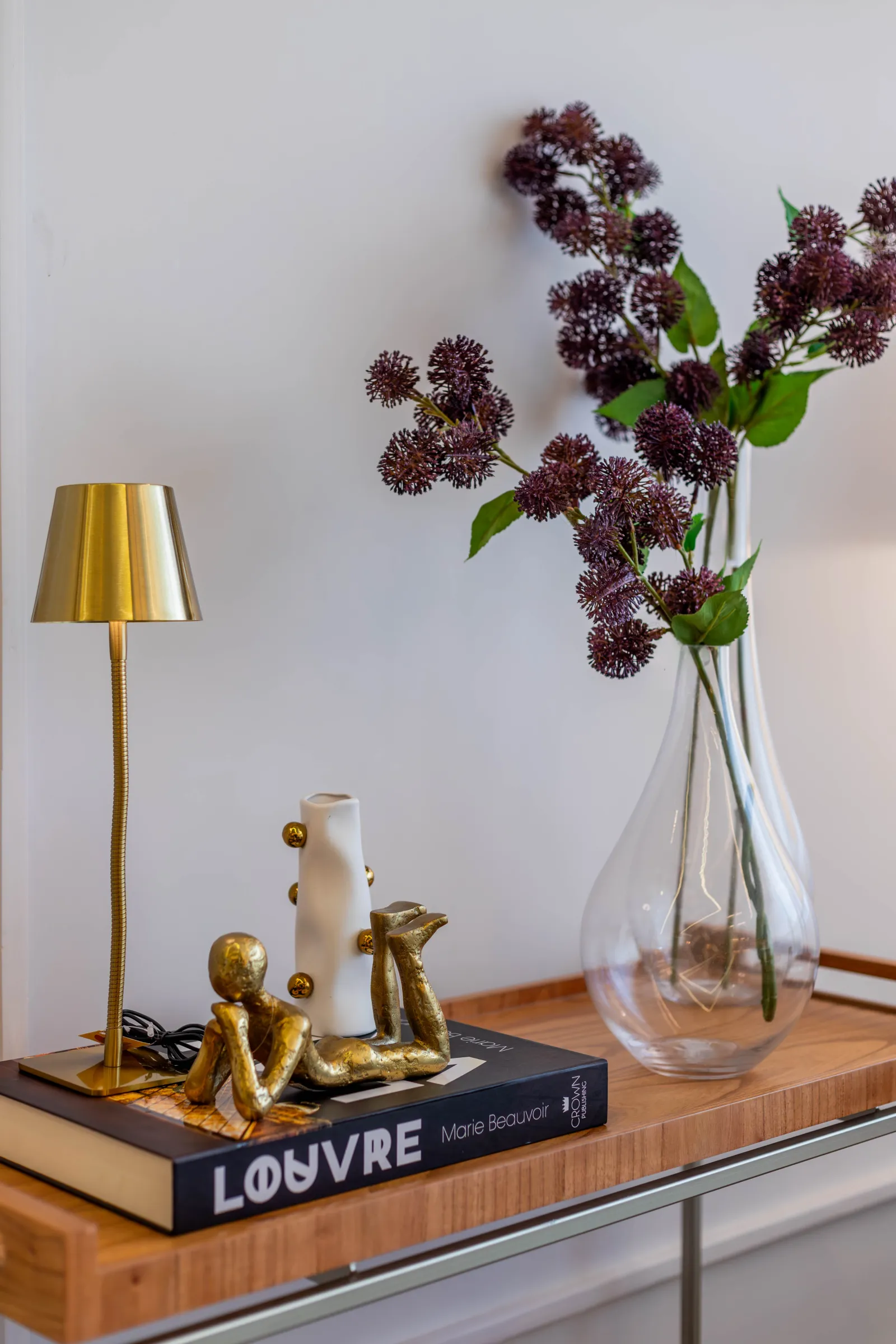 Detalhe decorativo do Residencial LV com aparador de madeira, abajur dourado, escultura decorativa e vaso de vidro com flores