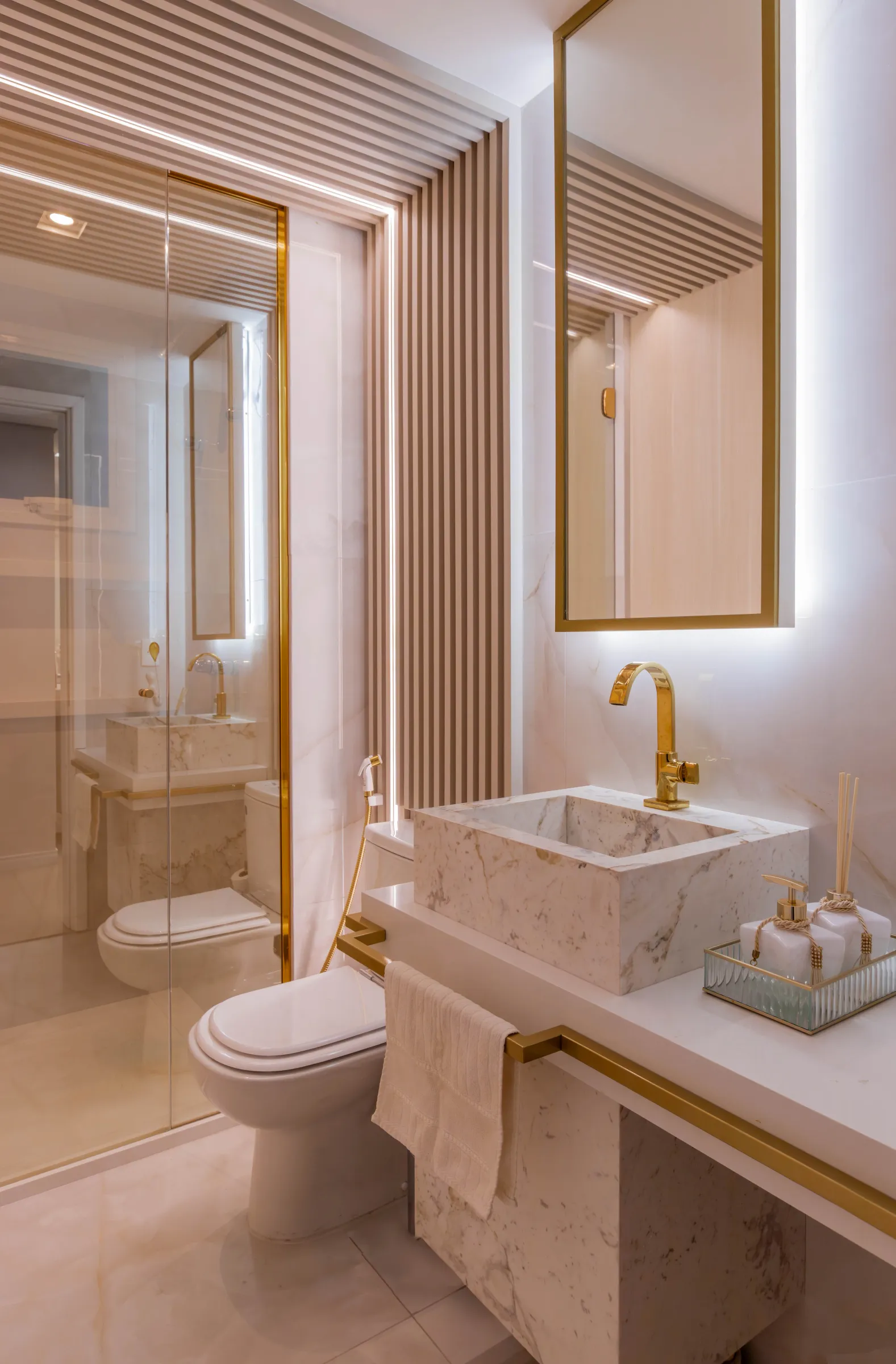 Lavabo do Residencial LV com cuba esculpida em mármore, torneira dourada, espelho com moldura dourada iluminada, painel ripado e box de vidro com perfil dourado