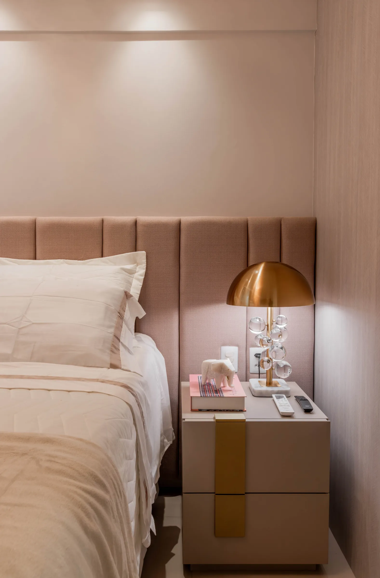 Quarto de casal do Residencial LV com cabeceira estofada capitonê rosé, roupa de cama branca e bege, criado-mudo com puxador dourado e abajur de cúpula dourada com base de esferas de cristal