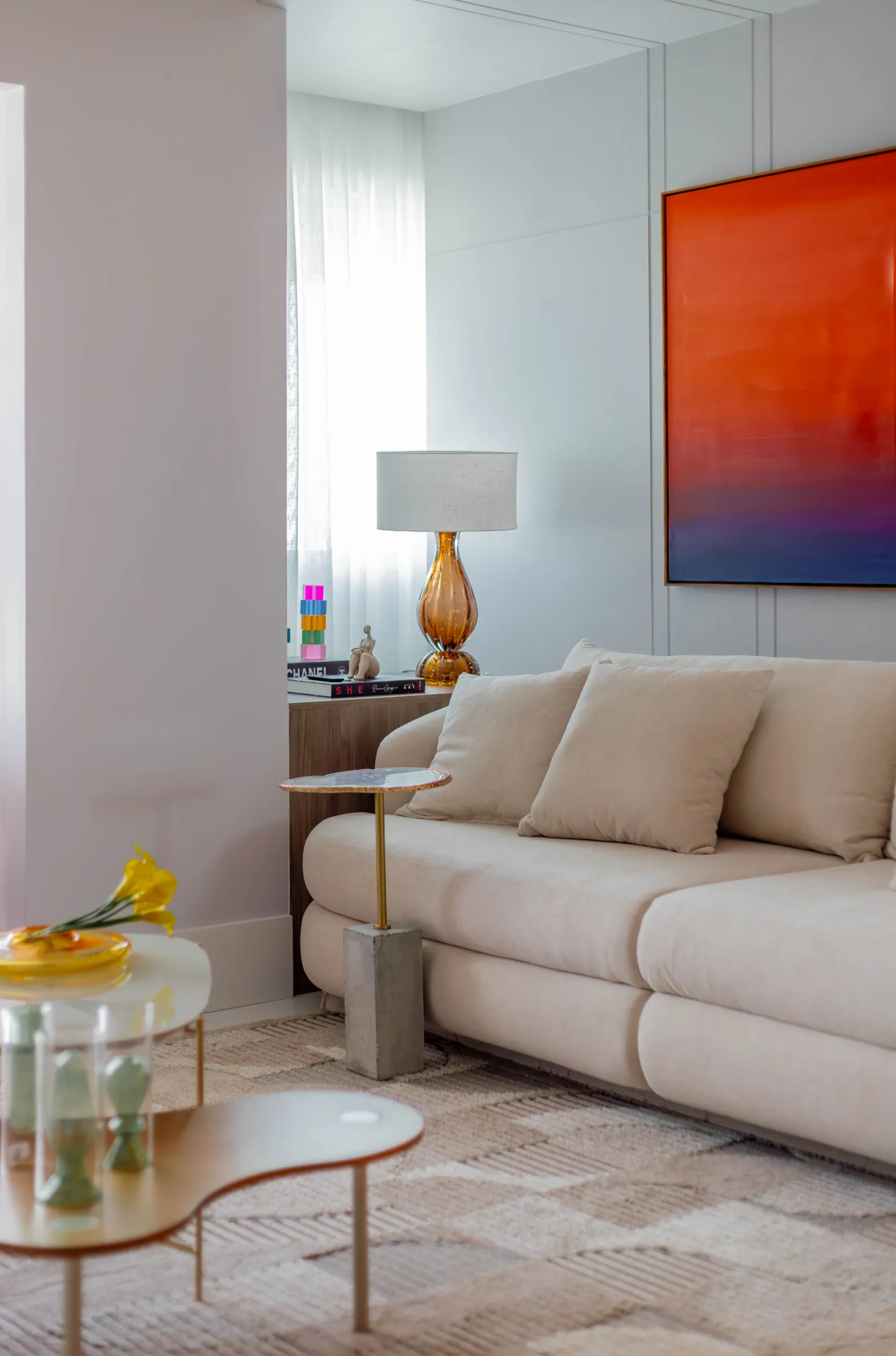 Sala de estar do Residencial ML com sofá curvo bege, grande quadro abstrato em laranja e azul, abajur de vidro âmbar sobre aparador de madeira e mesa de centro orgânica
