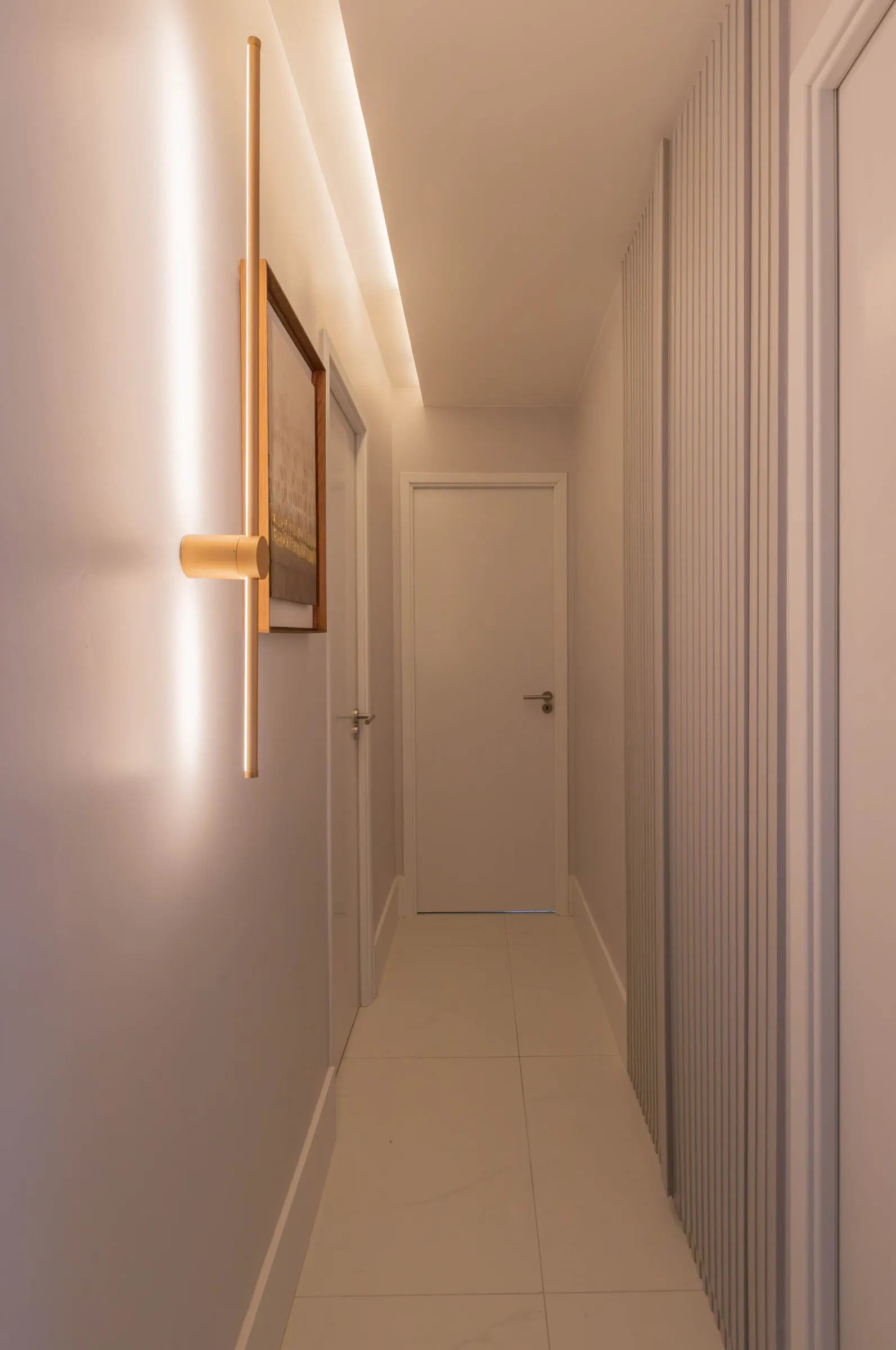 Corredor do Residencial ML com arandela dourada vertical, quadro emoldurado, painel ripado branco e piso porcelanato claro