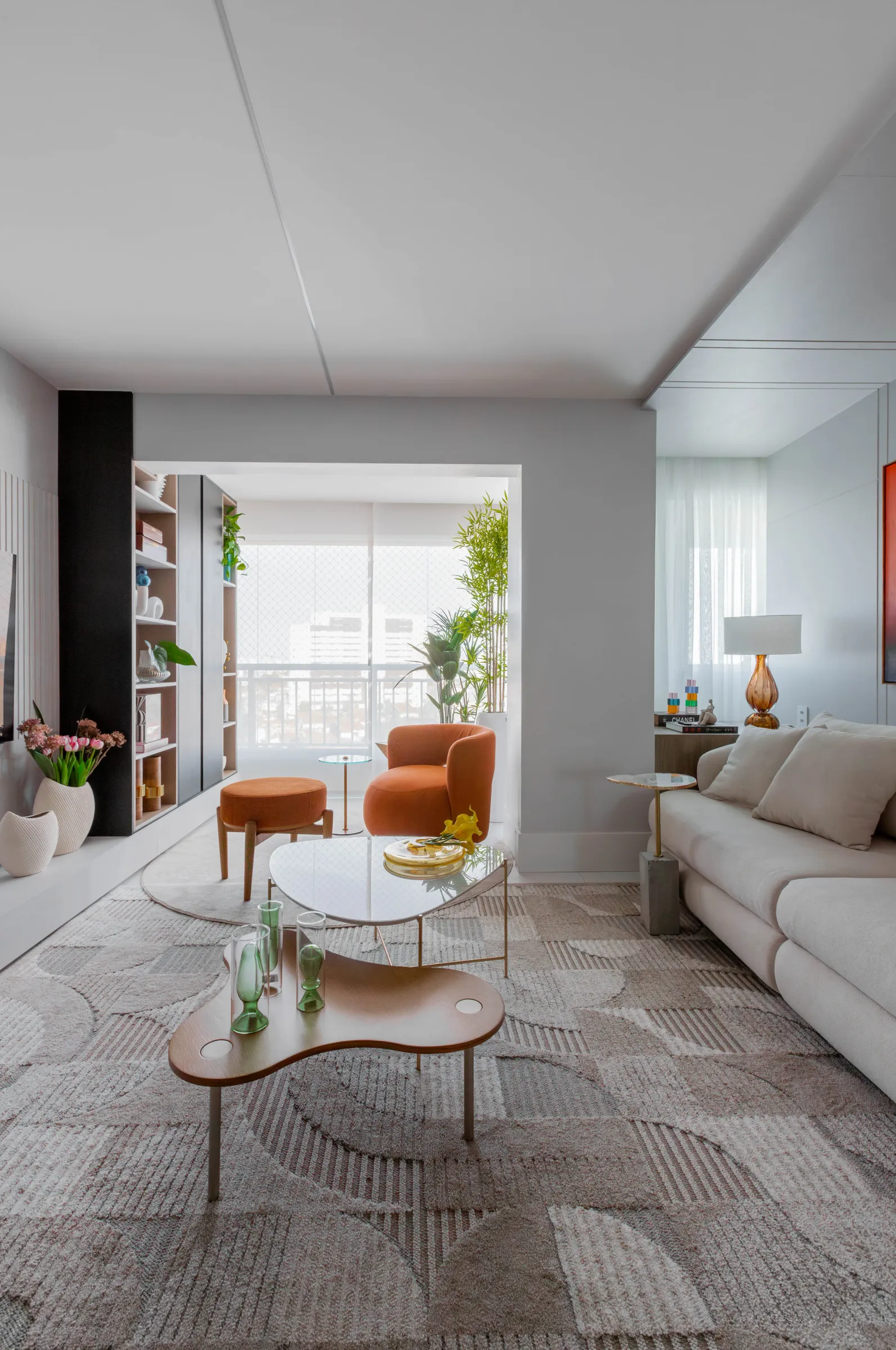 Sala de estar do Residencial ML com sofá bege, poltrona e puff terracota, estante de nichos, mesas de centro orgânicas e varanda com plantas ao fundo