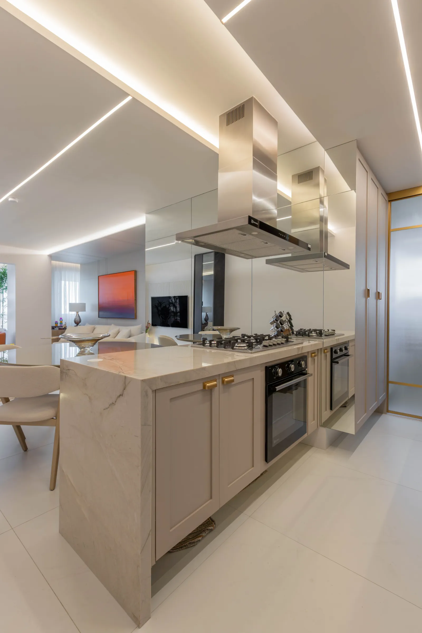 Cozinha do Residencial ML com bancada de mármore, coifa de inox, cooktop e forno embutidos, marcenaria off-white com puxadores dourados e painel espelhado