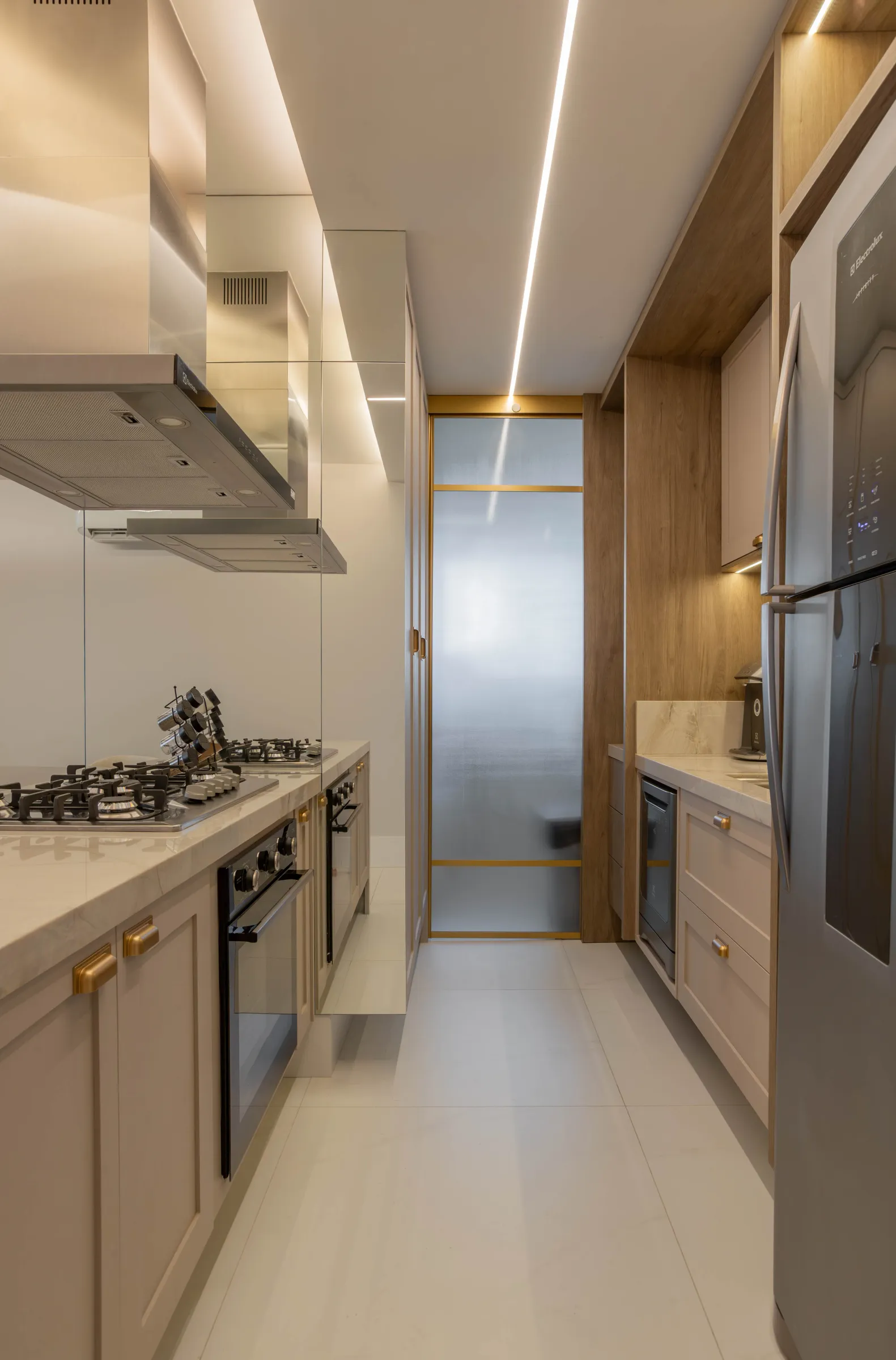 Cozinha do Residencial ML em layout corredor, com cooktop, coifa de inox, fornos embutidos, geladeira inox e marcenaria clara com puxadores dourados e porta de vidro para a área de serviço