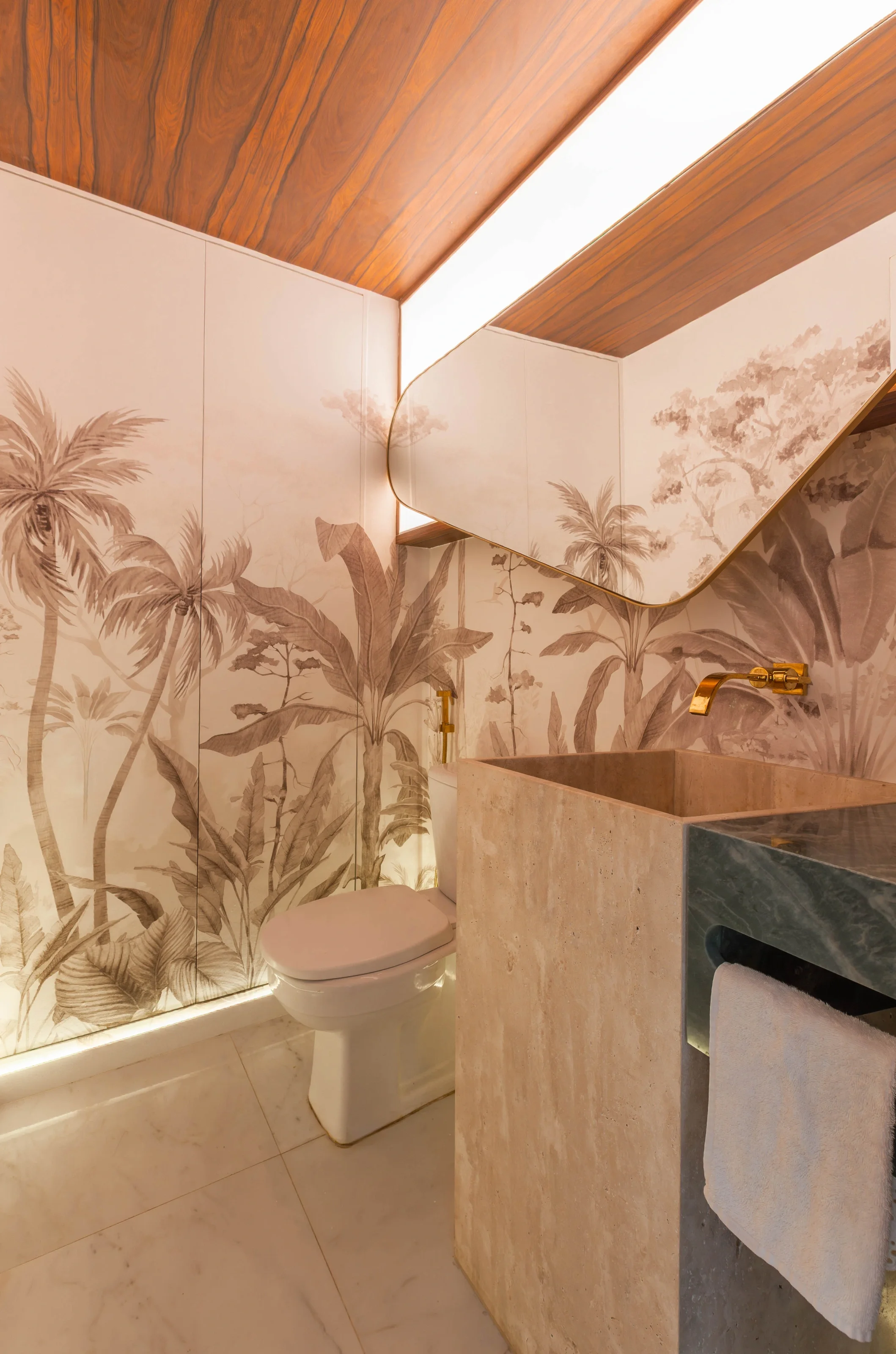 Lavabo do Residencial PL com papel de parede tropical de palmeiras em tons sépia, cuba esculpida em pedra, torneira dourada de parede, espelho orgânico e forro amadeirado