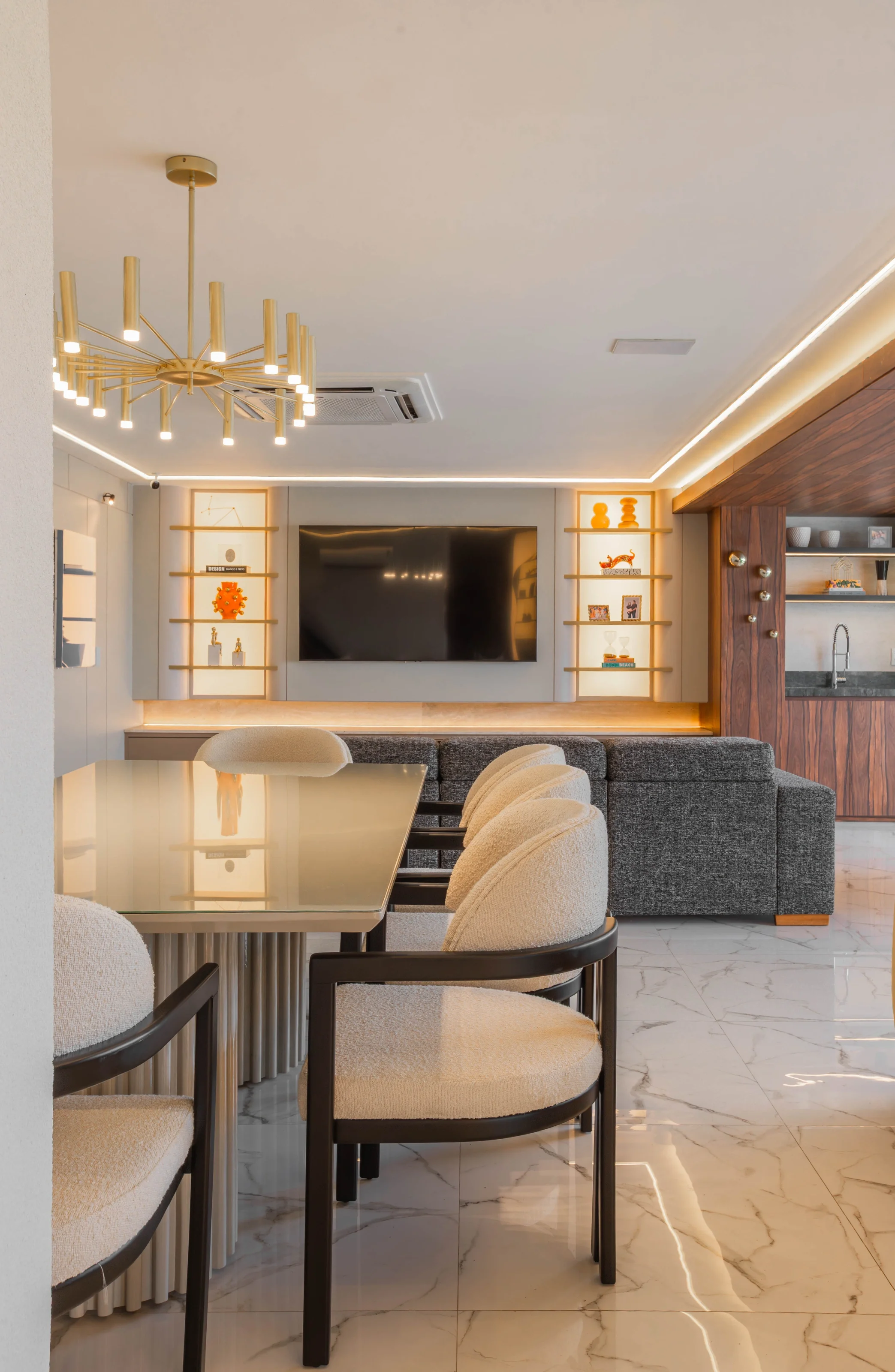 Sala de estar do Residencial PL com painel de TV e nichos iluminados, sofá cinza, marcenaria amadeirada com bar e mesa de jantar com cadeiras de bouclé