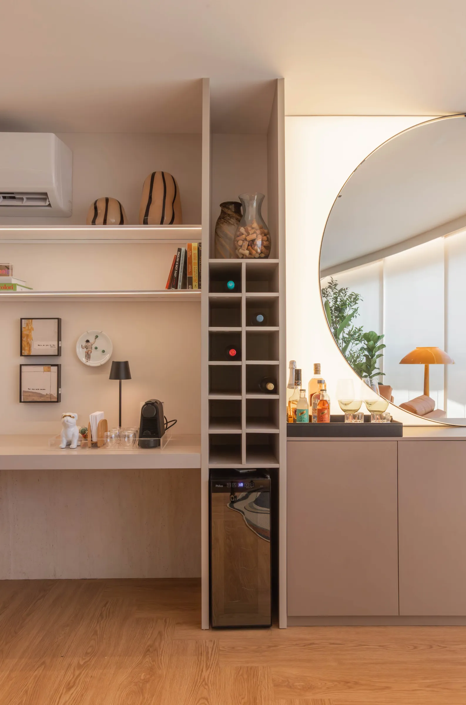 Detalhe do bar do Residencial RC com adega de garrafas embutida, frigobar, cafeteira, espelho redondo e bandeja com bebidas e taças, ao lado de bancada com quadros