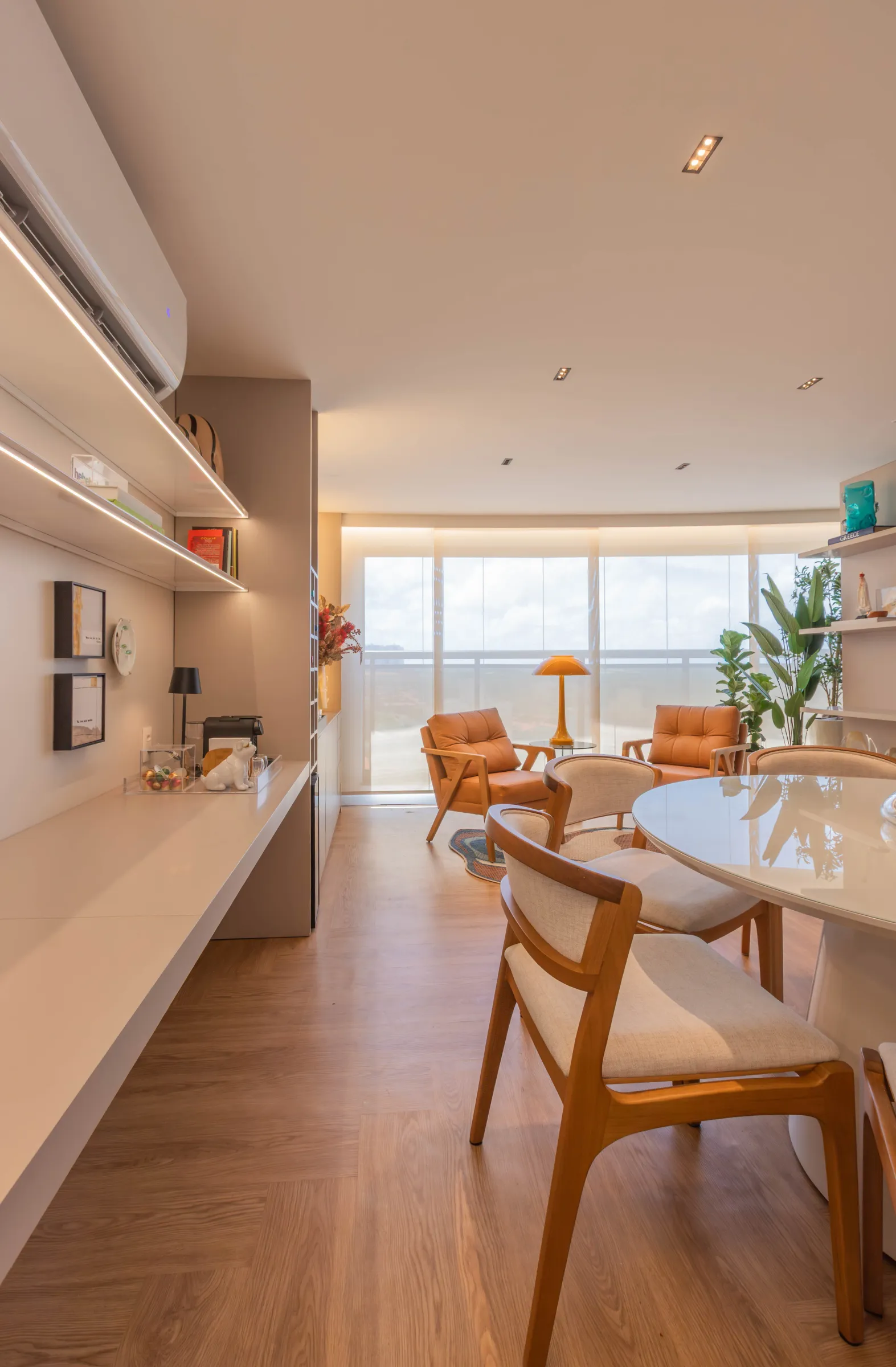 Sala de jantar do Residencial RC com mesa redonda branca e cadeiras de madeira estofadas, poltronas de couro caramelo junto à varanda e aparador branco com prateleiras iluminadas