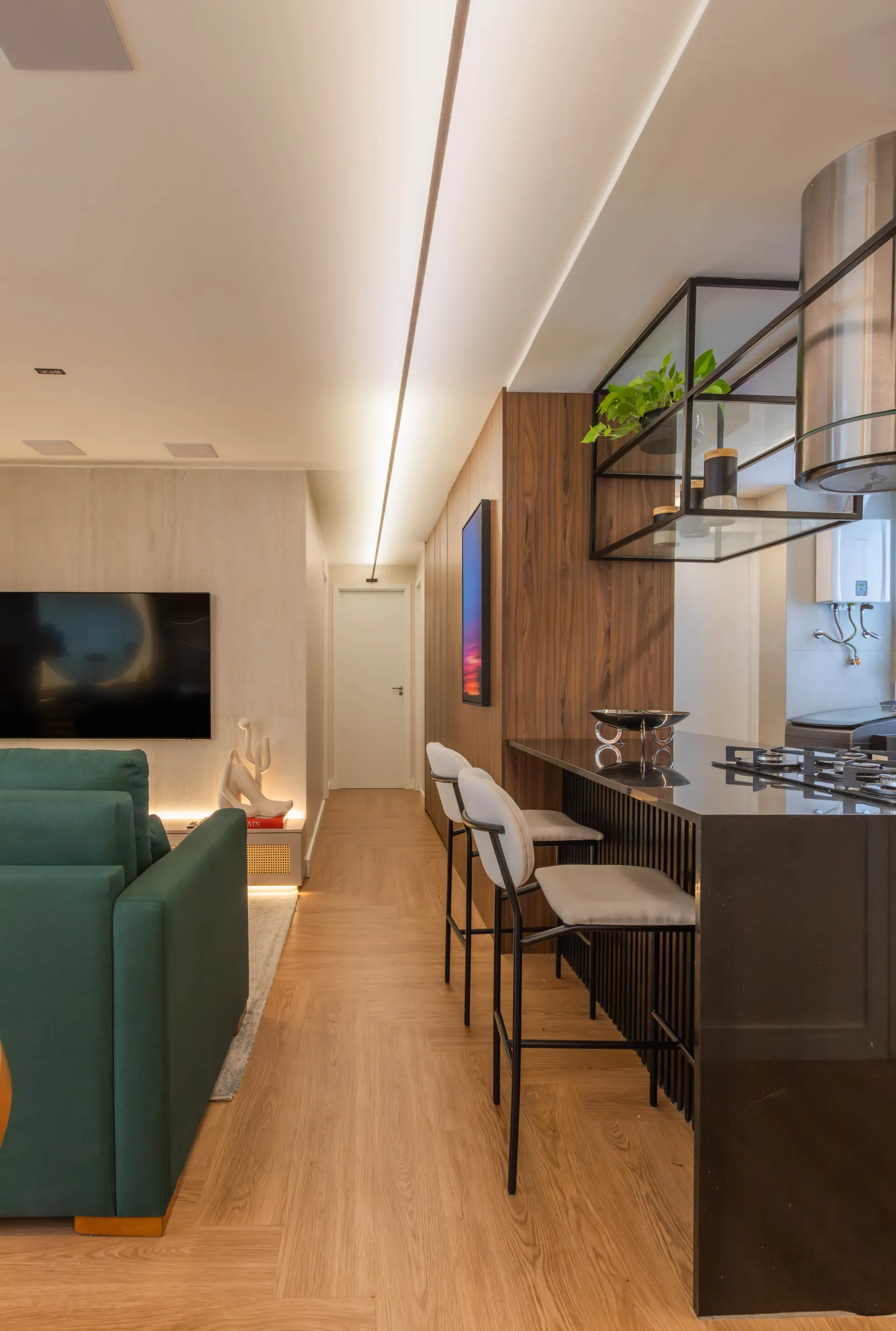 Living do Residencial RC com sofá verde, painel de TV em mármore e integração com a cozinha americana de bancada preta, banquetas e coifa de inox