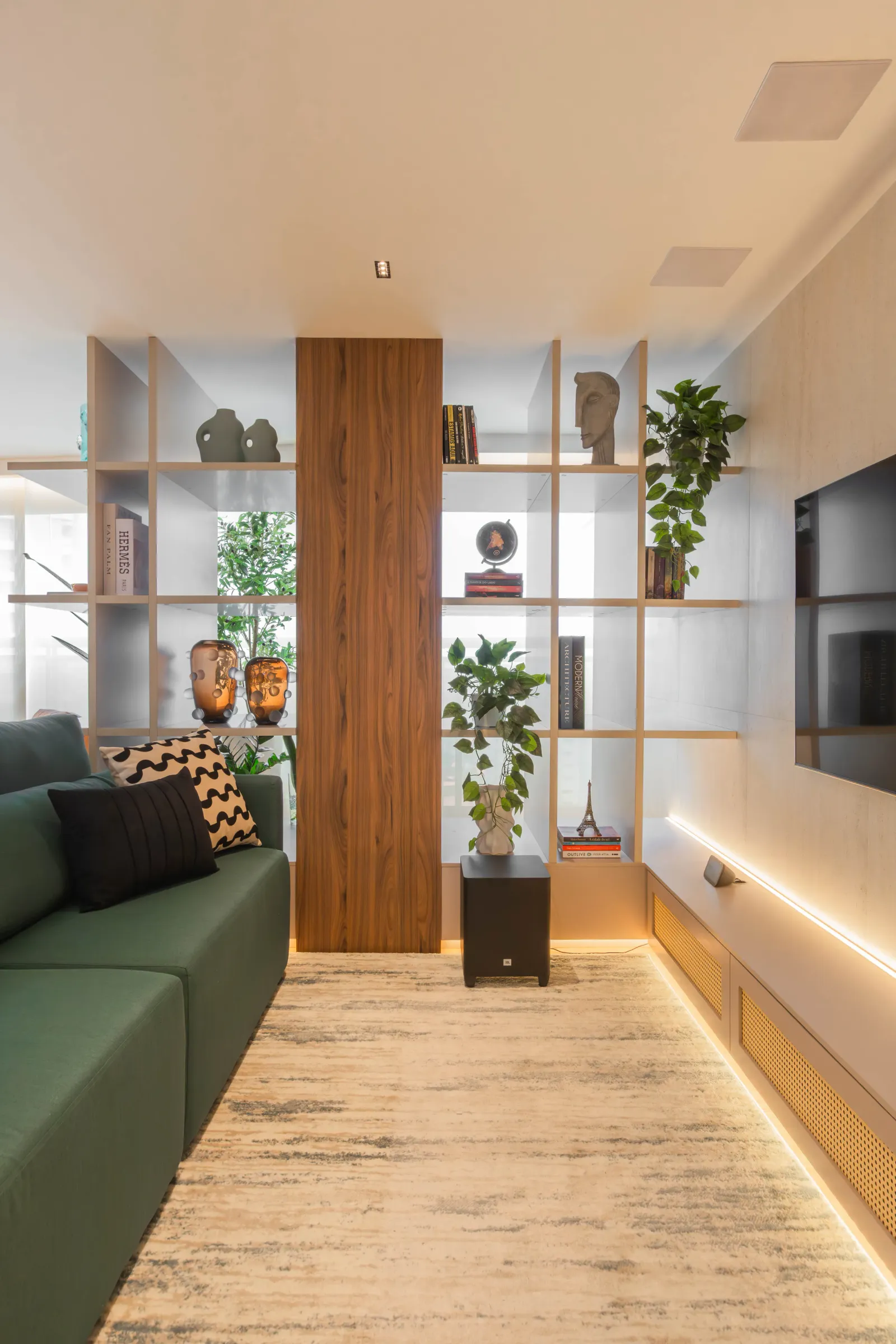 Sala de TV do Residencial RC com sofá verde, estante vazada bege com plantas e objetos decorativos, coluna de madeira, painel de TV e tapete claro