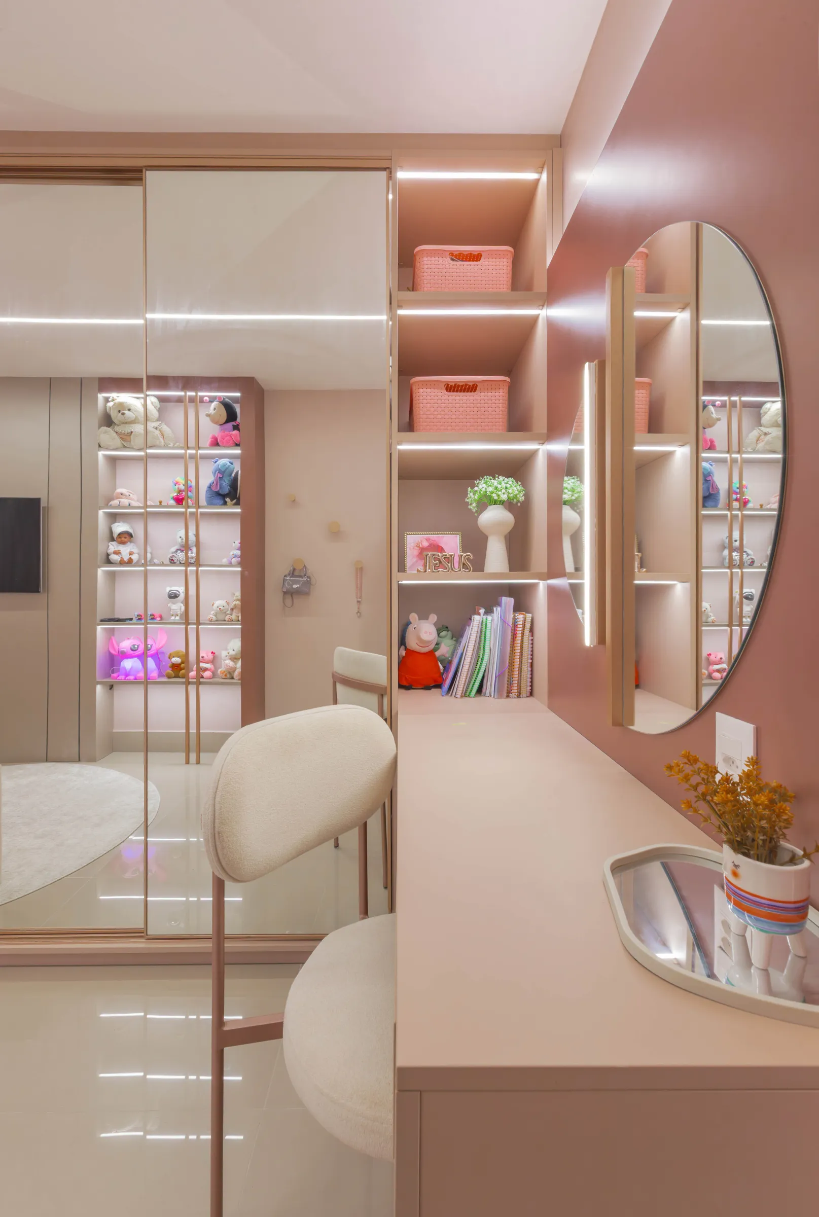 Quarto infantil do Residencial TN em tons de rosé, com bancada de estudos, prateleiras iluminadas expondo pelúcias, espelho oval e guarda-roupa espelhado