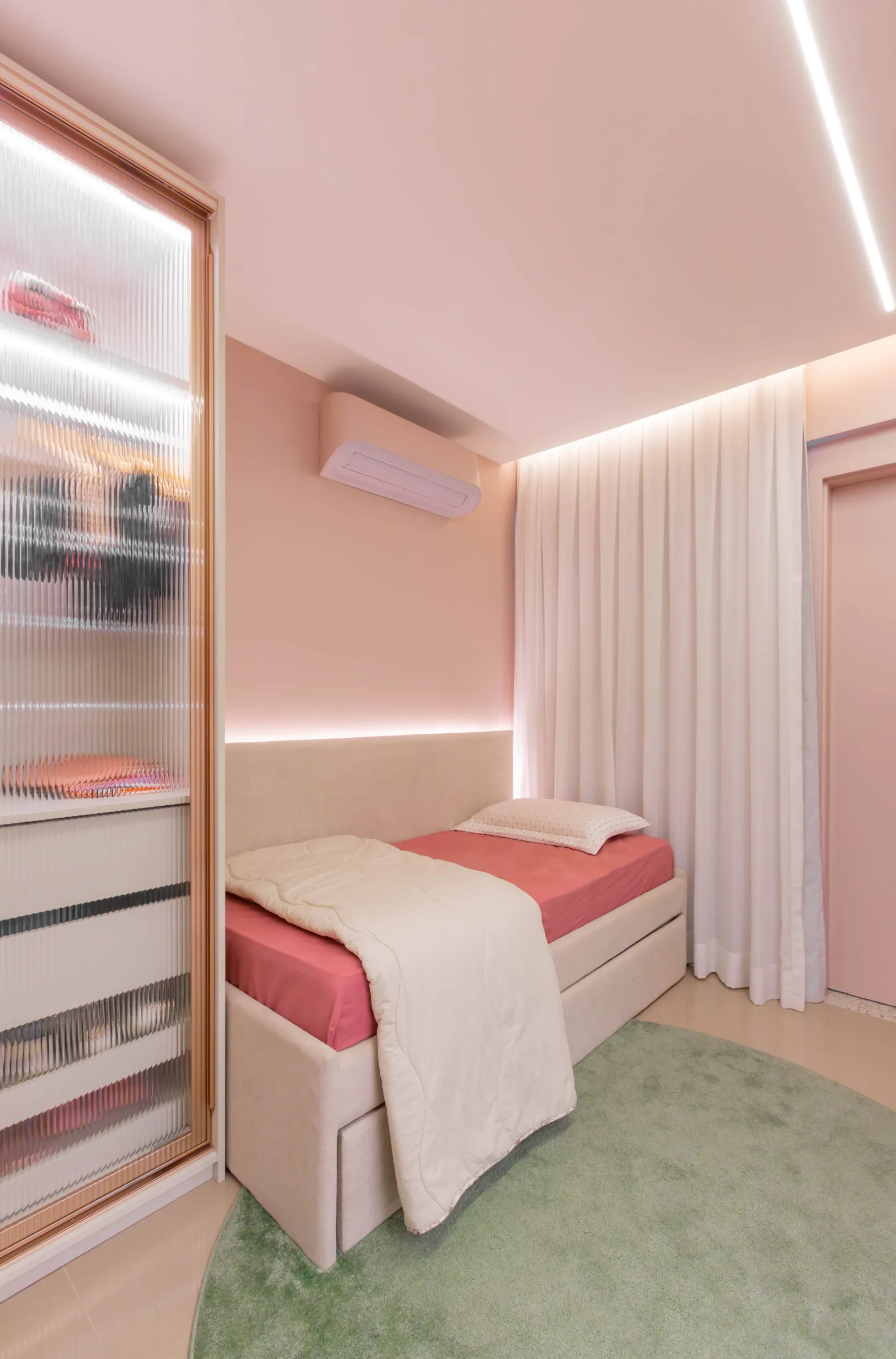 Quarto de solteiro do Residencial TN com parede rosé, cama com roupa de cama em rosa e terracota, tapete redondo verde e guarda-roupa de vidro canelado