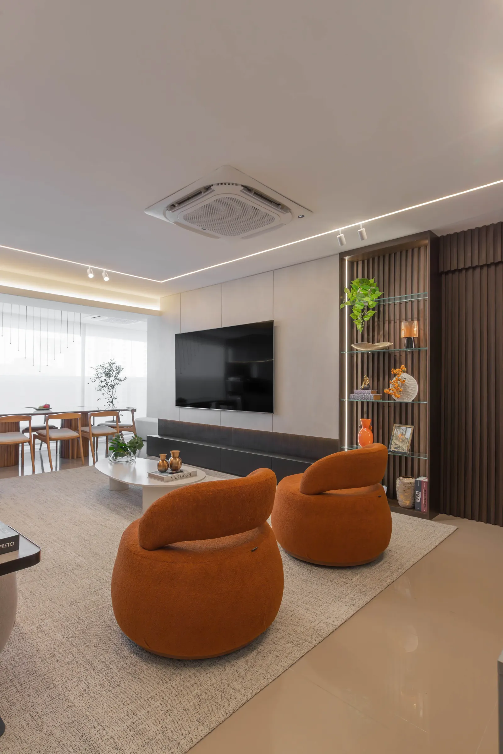 Living do Residencial TN com duas poltronas laranja arredondadas, painel de TV, estante ripada de madeira com prateleiras de vidro iluminadas e integração com a sala de jantar