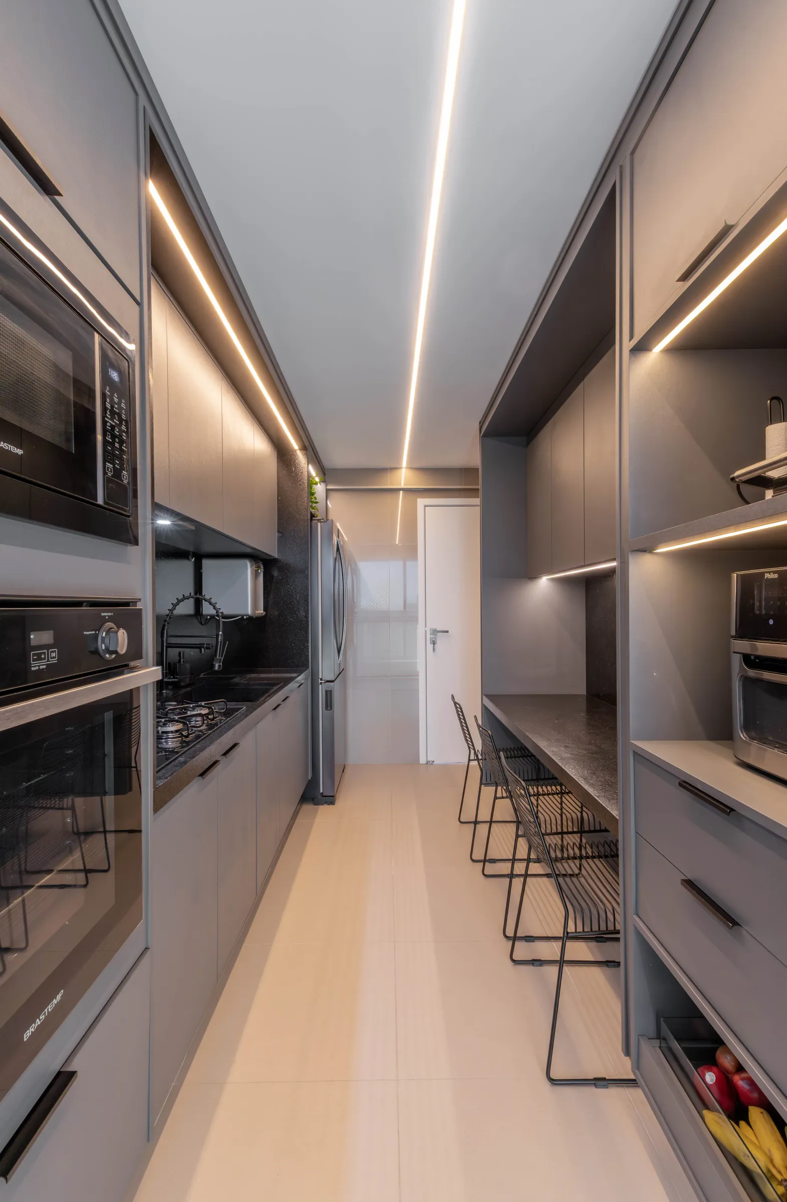 Cozinha planejada do Residencial TN em layout corredor, com marcenaria cinza, fornos e micro-ondas embutidos, cooktop, geladeira inox e bancada com banquetas de fio preto