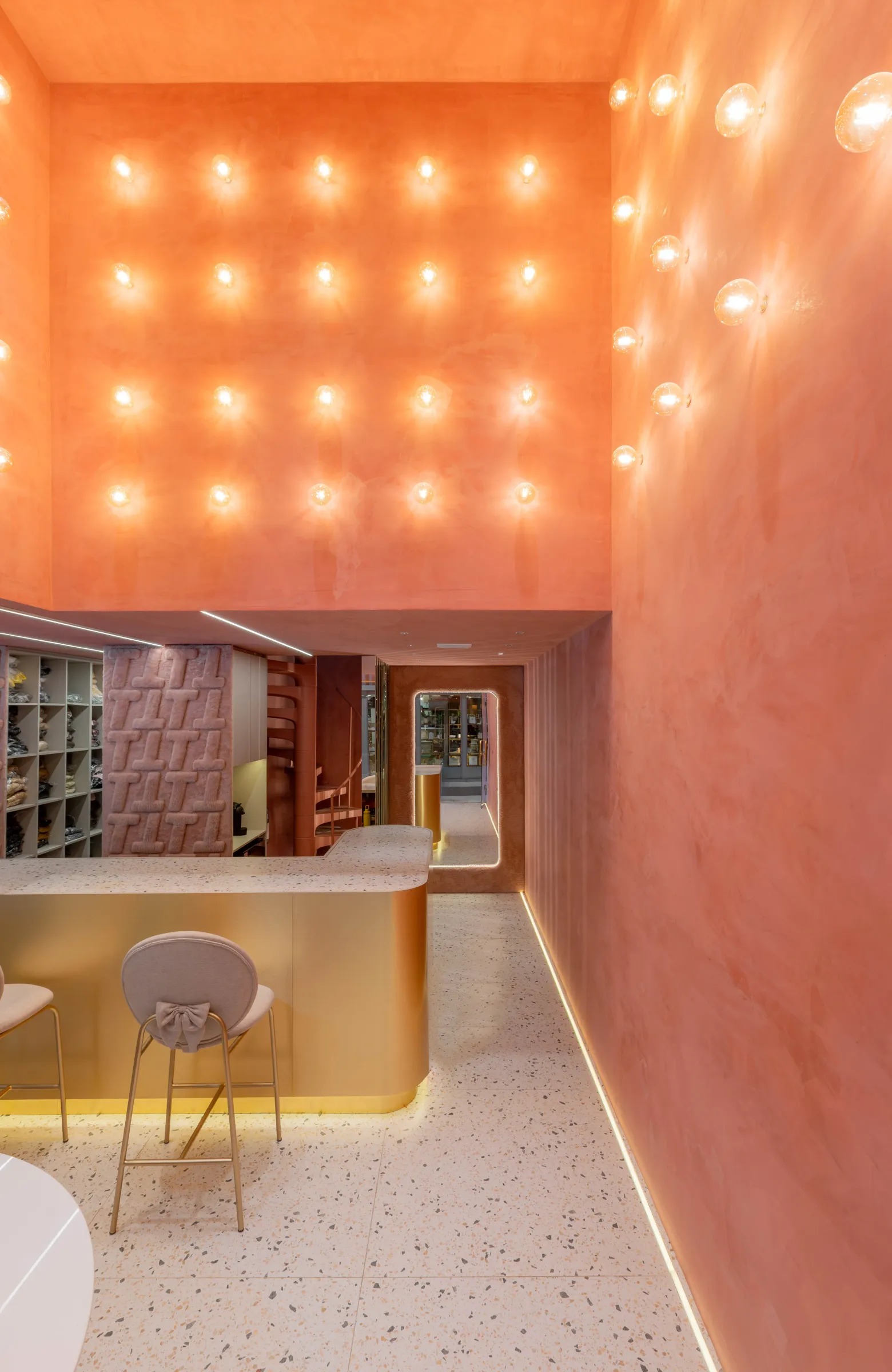 Interior da loja Teca com parede alta terracota pontuada por lâmpadas acesas em grade, balcão de terrazzo e latão com banquetas rosa de laço, painel de pelúcia com monograma T e corredor iluminado ao fundo